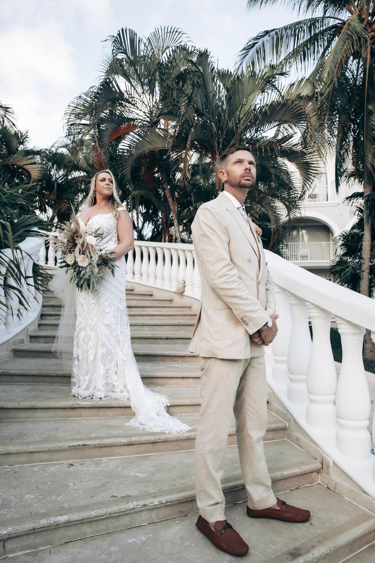 Bride and groom posing on white staircase with palm trees at tropical wedding venue.
