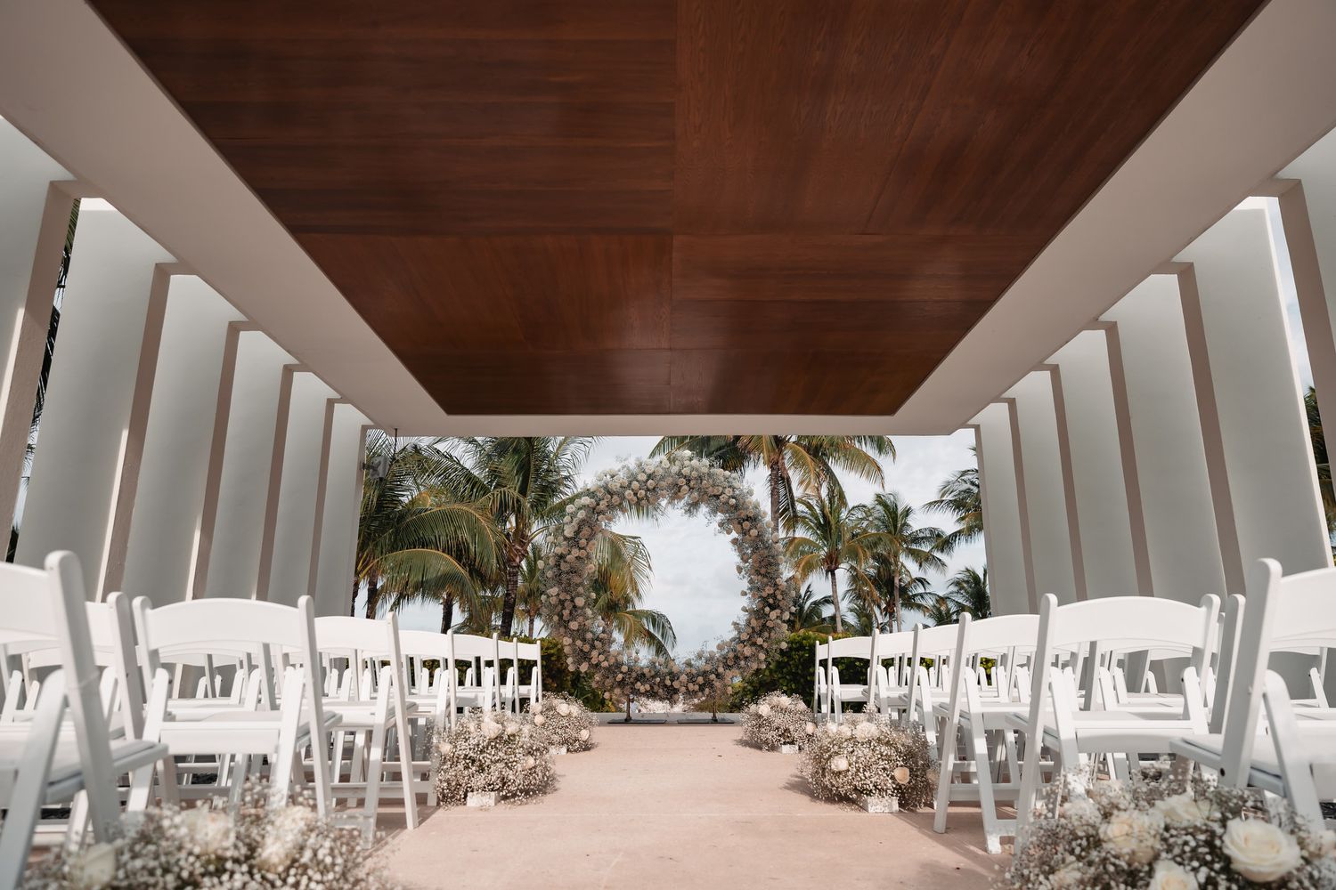 Elegant outdoor wedding ceremony aisle with white chairs and a circular floral arch framing tropical palm trees.