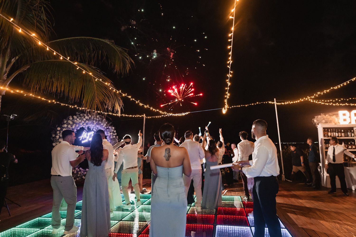 Wedding guests celebrate with sparklers on colorful LED dance floor as fireworks light up night sky.