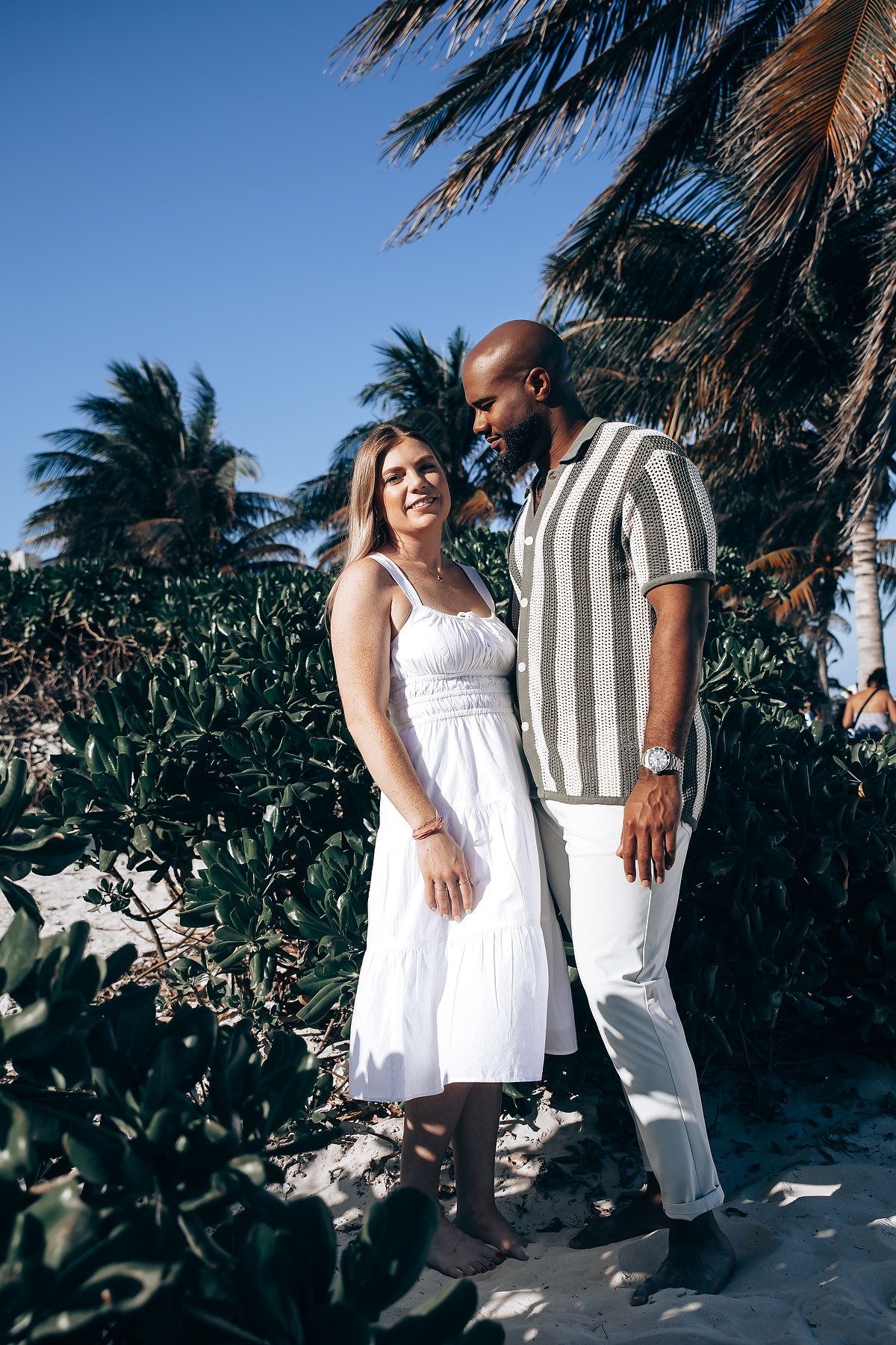Interracial couple in white dress and striped shirt standing together on a tropical beach with palms.