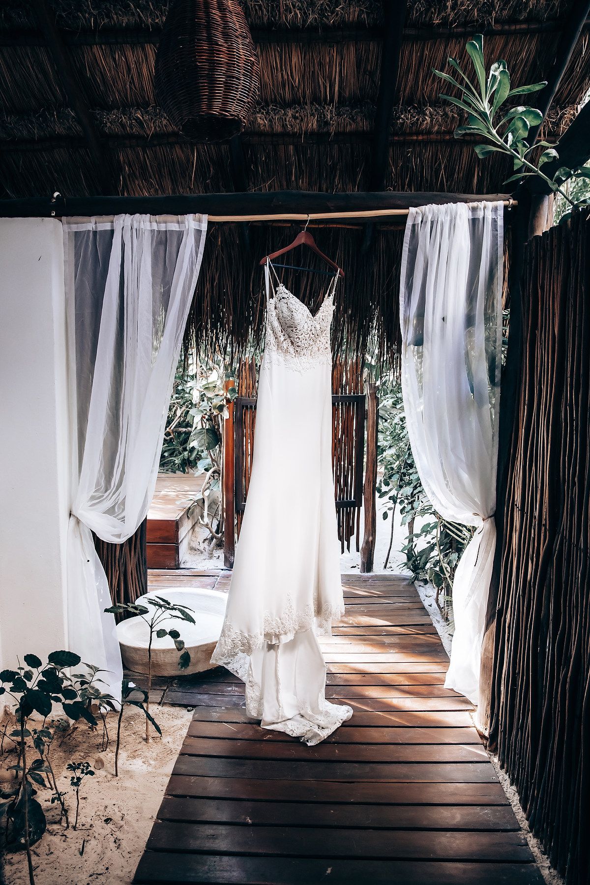 A white wedding dress hangs in a doorway decorated with sheer curtains and greenery.