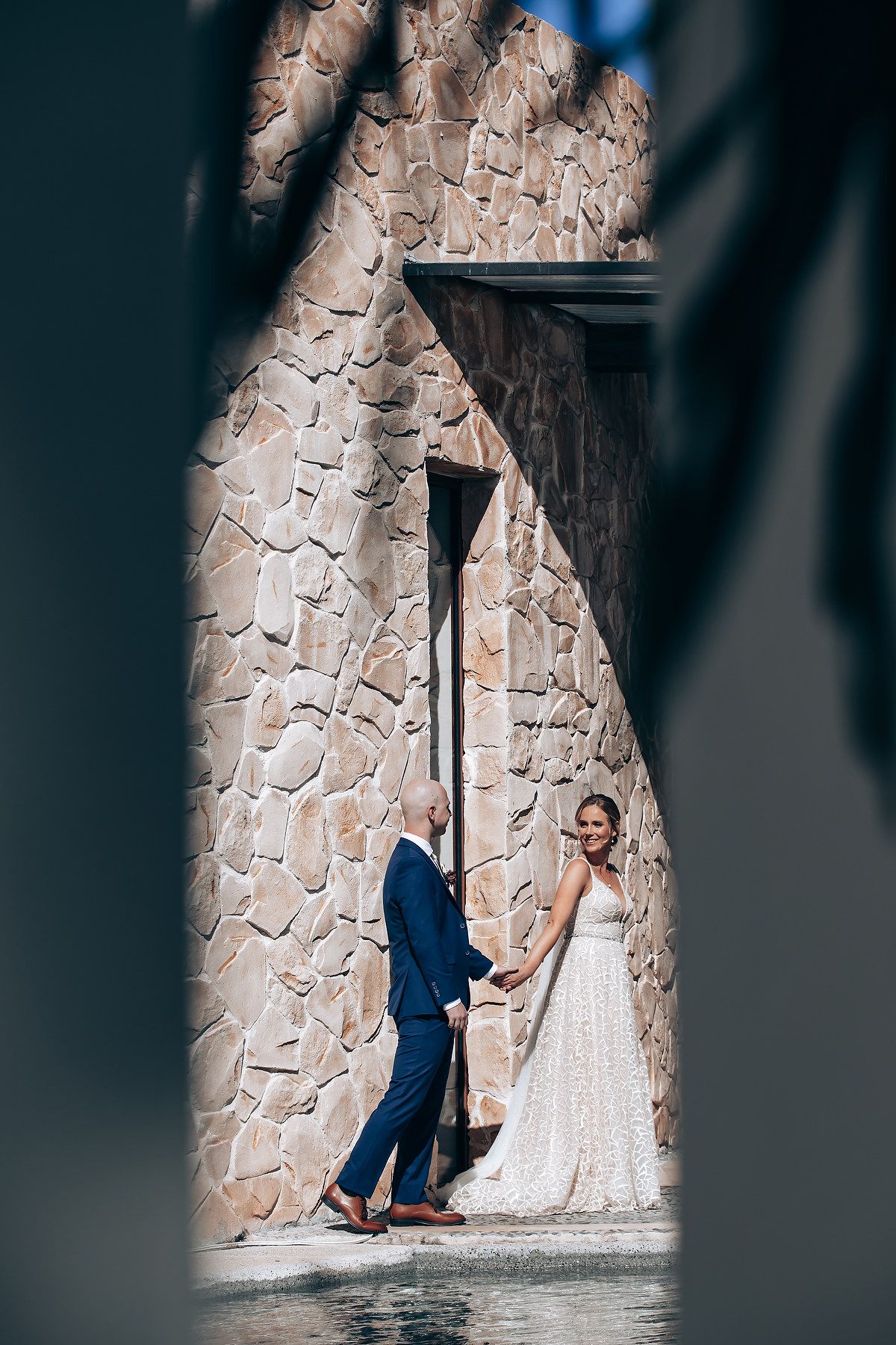 A bride and groom walk together through a stone archway, the bride wearing a long, flowing wedding dress.