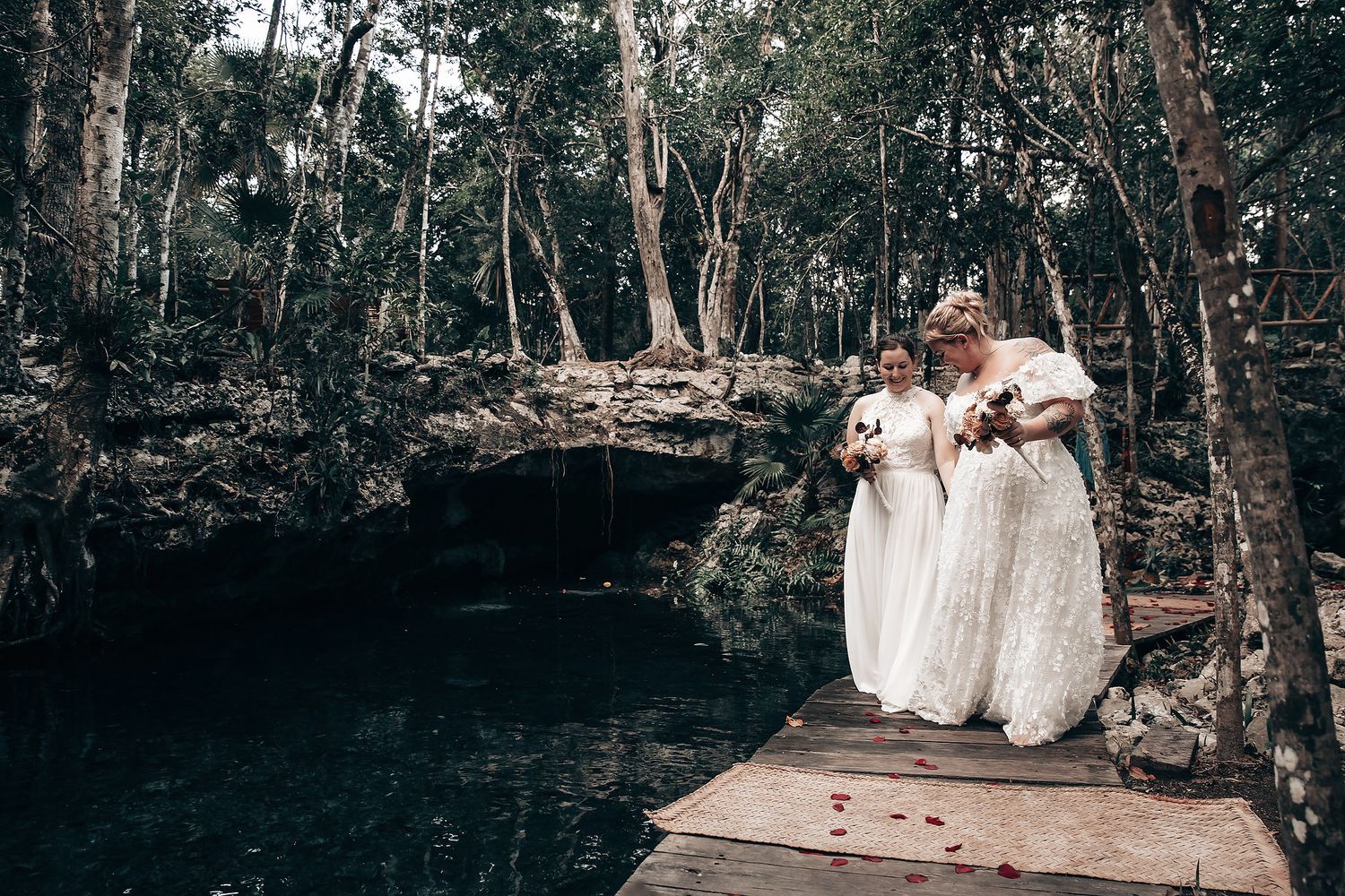 Two women in white wedding dresses standing on a wooden dock in a natural setting.