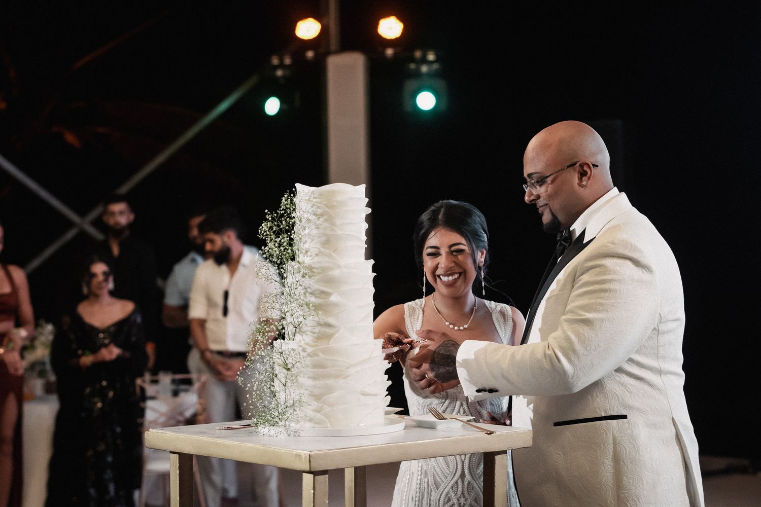 Smiling bride and groom cut their elegant multi-tiered white wedding cake adorned with delicate floral decorations.