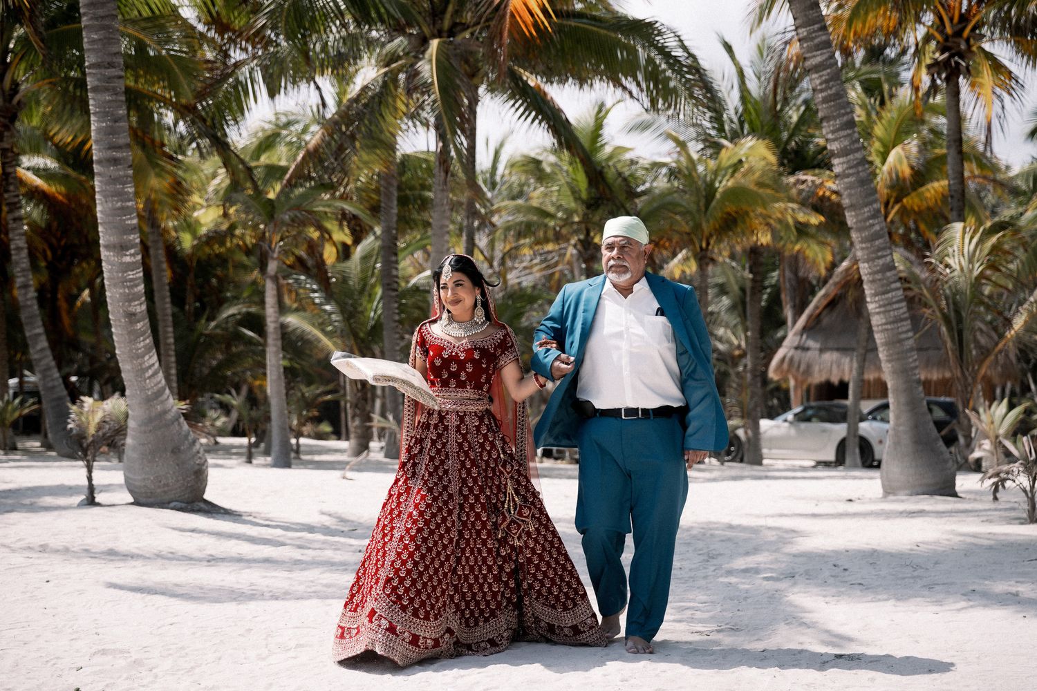 Bride in red lehenga walks with elder on sandy beach surrounded by palm trees.
