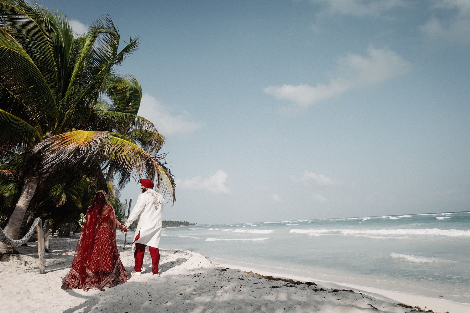 Indian couple in traditional red and white wedding attire holding hands on tropical beach with palm trees.