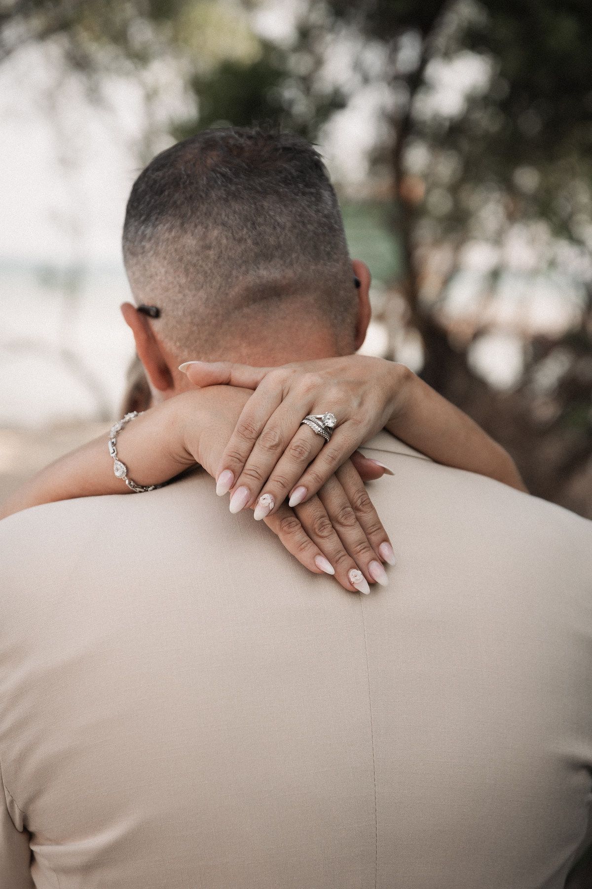 Close-up of bride's hand with wedding ring set resting on groom's shoulder during beach wedding.