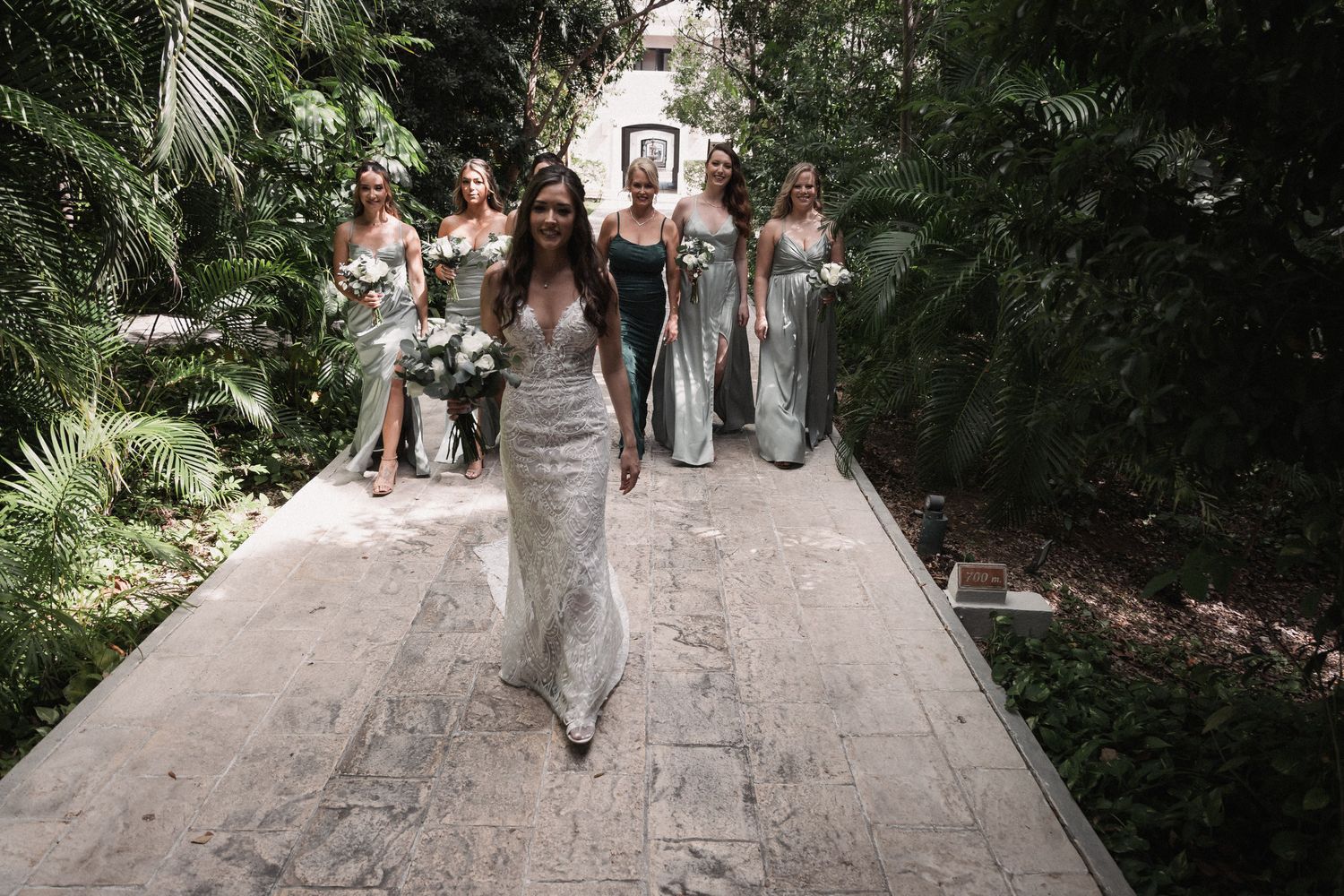 Bride in lace gown leads bridesmaids in silver dresses down tropical garden pathway before wedding ceremony.