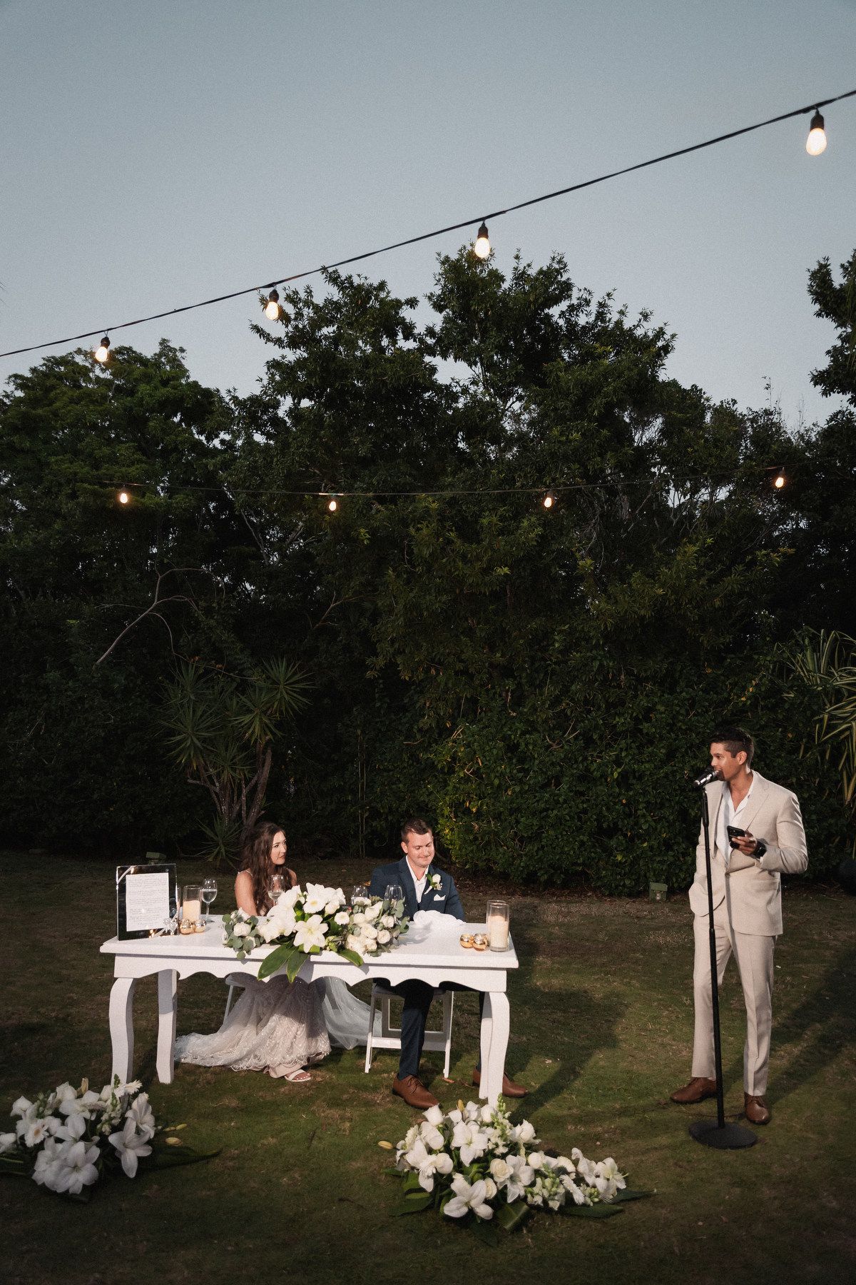 Wedding couple sits at sweetheart table while groomsman delivers speech at outdoor evening reception.