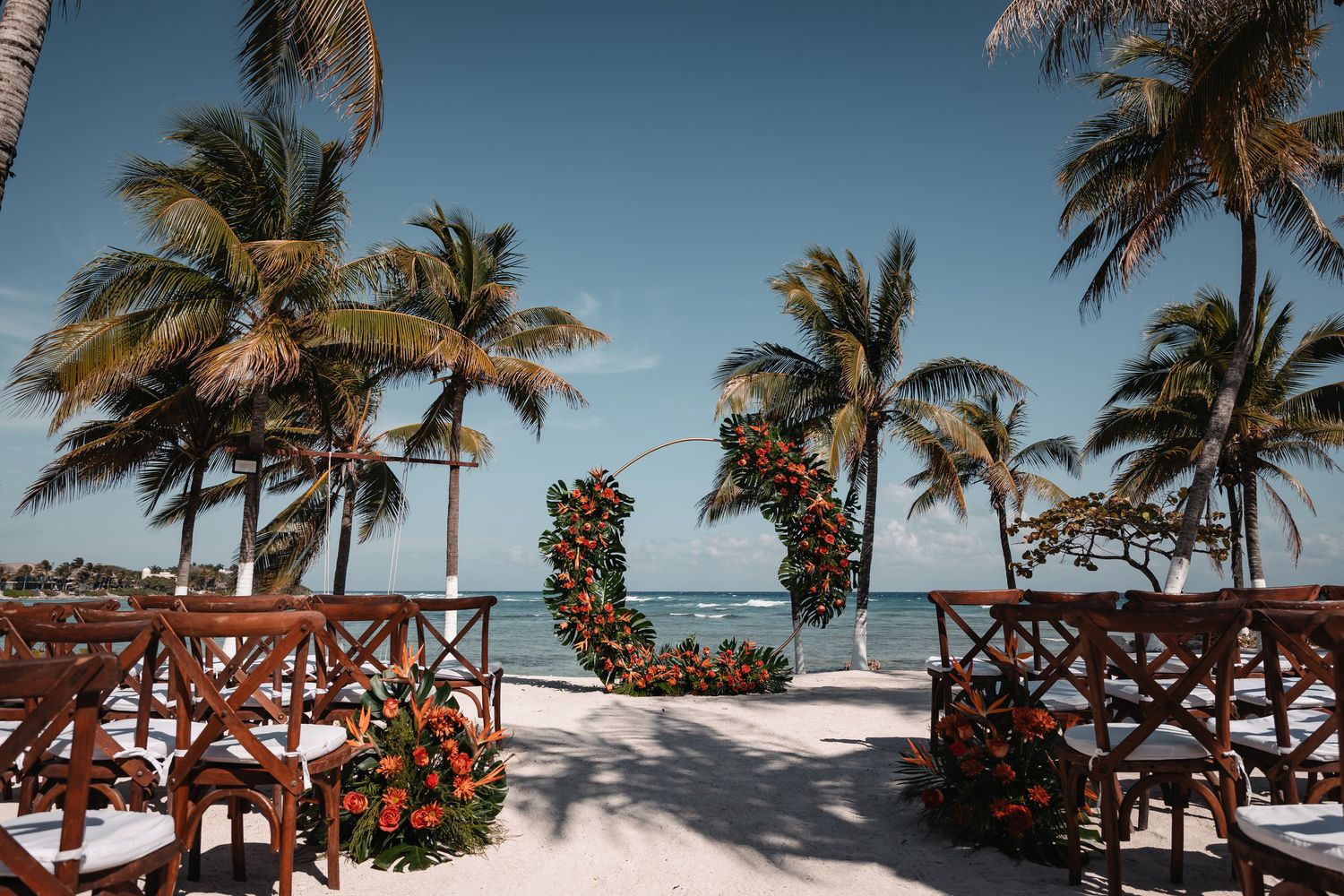 Beach wedding setup with floral arch, wooden chairs, and palm trees overlooking a turquoise ocean on a sunny day.