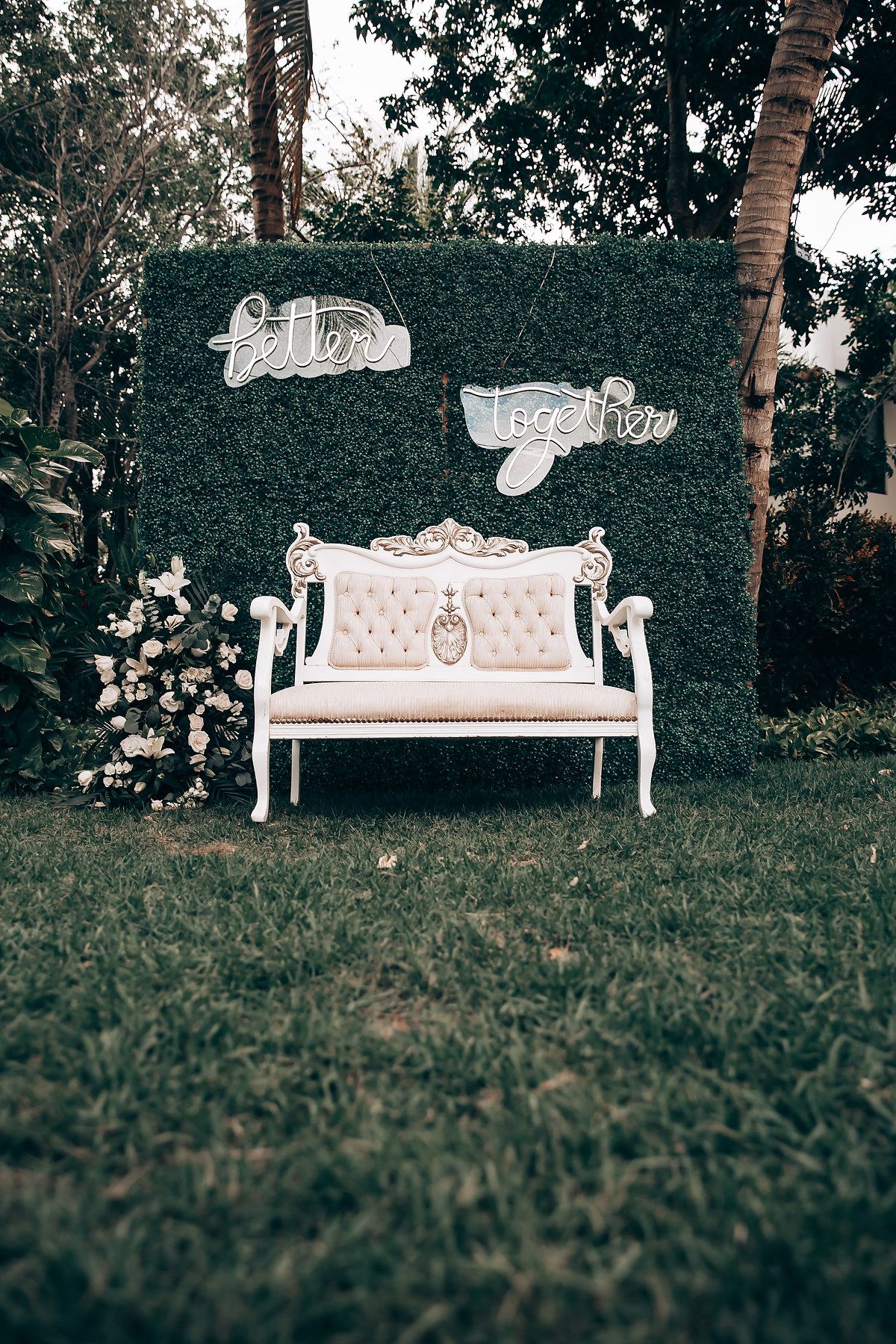 White vintage bench against a green hedge wall with decorative signage for a wedding photo backdrop.