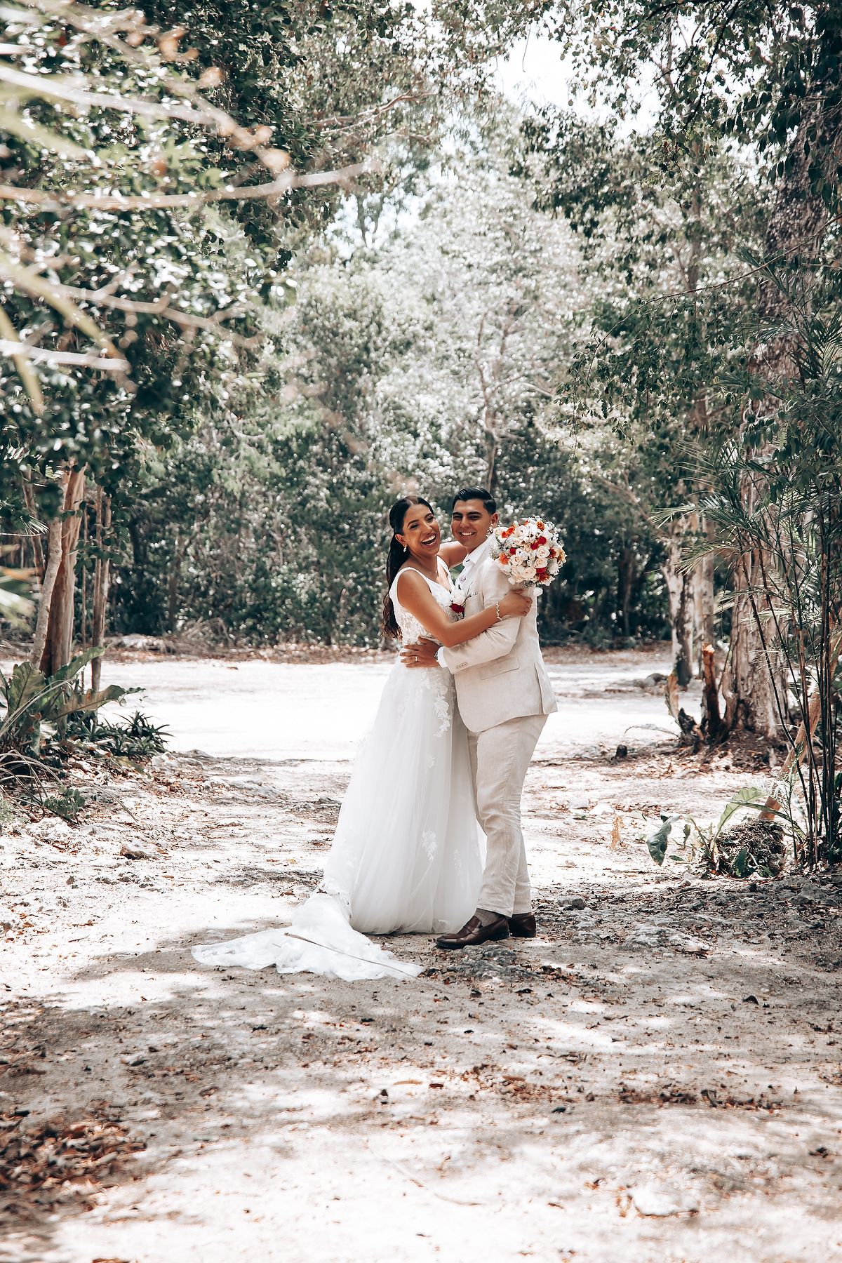 Newlyweds share intimate moment on forest path in white and beige attire.