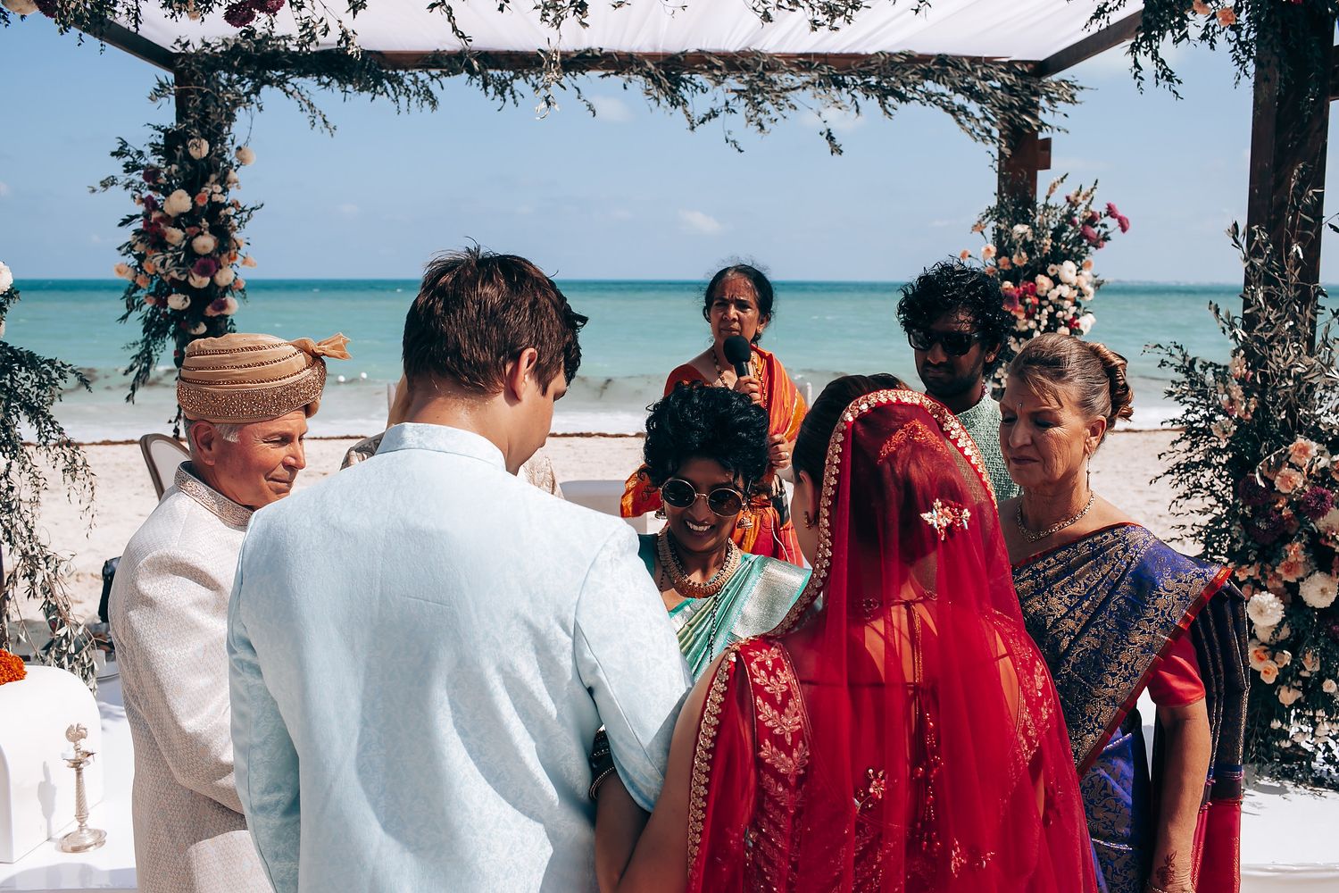 A beachside wedding ceremony takes place under an arch decorated with flowers and greenery.
