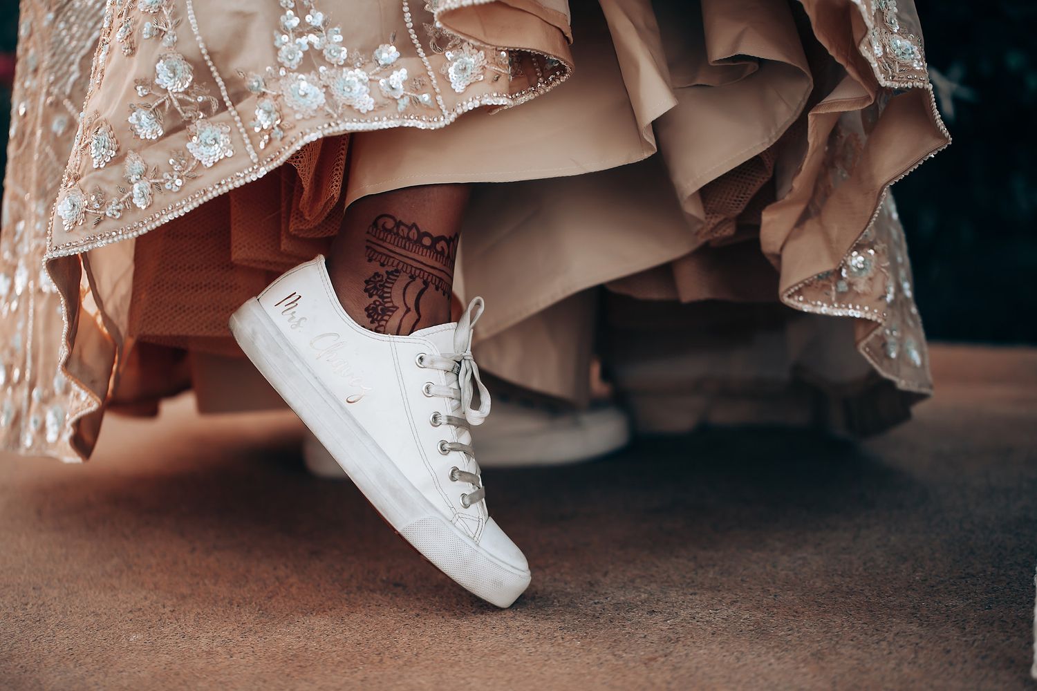 Close-up detail shot of ornate wedding dress hem and white shoes on dark floor.