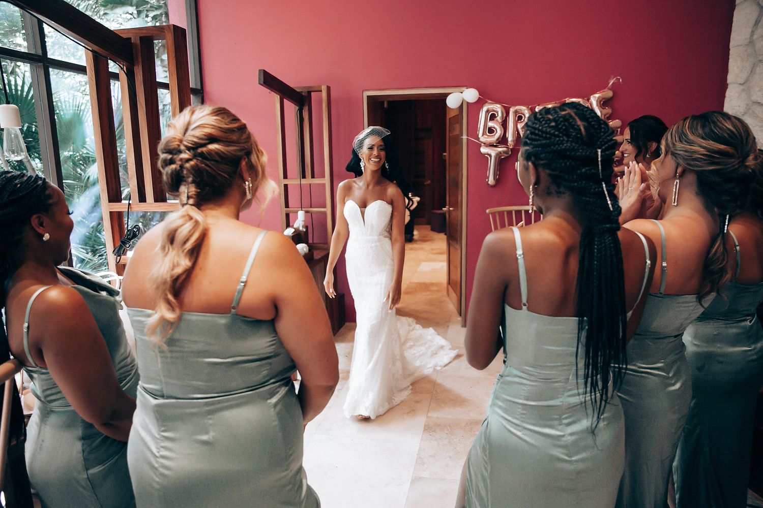 A bride in a white dress stands centered while bridesmaids in sage dresses surround her.