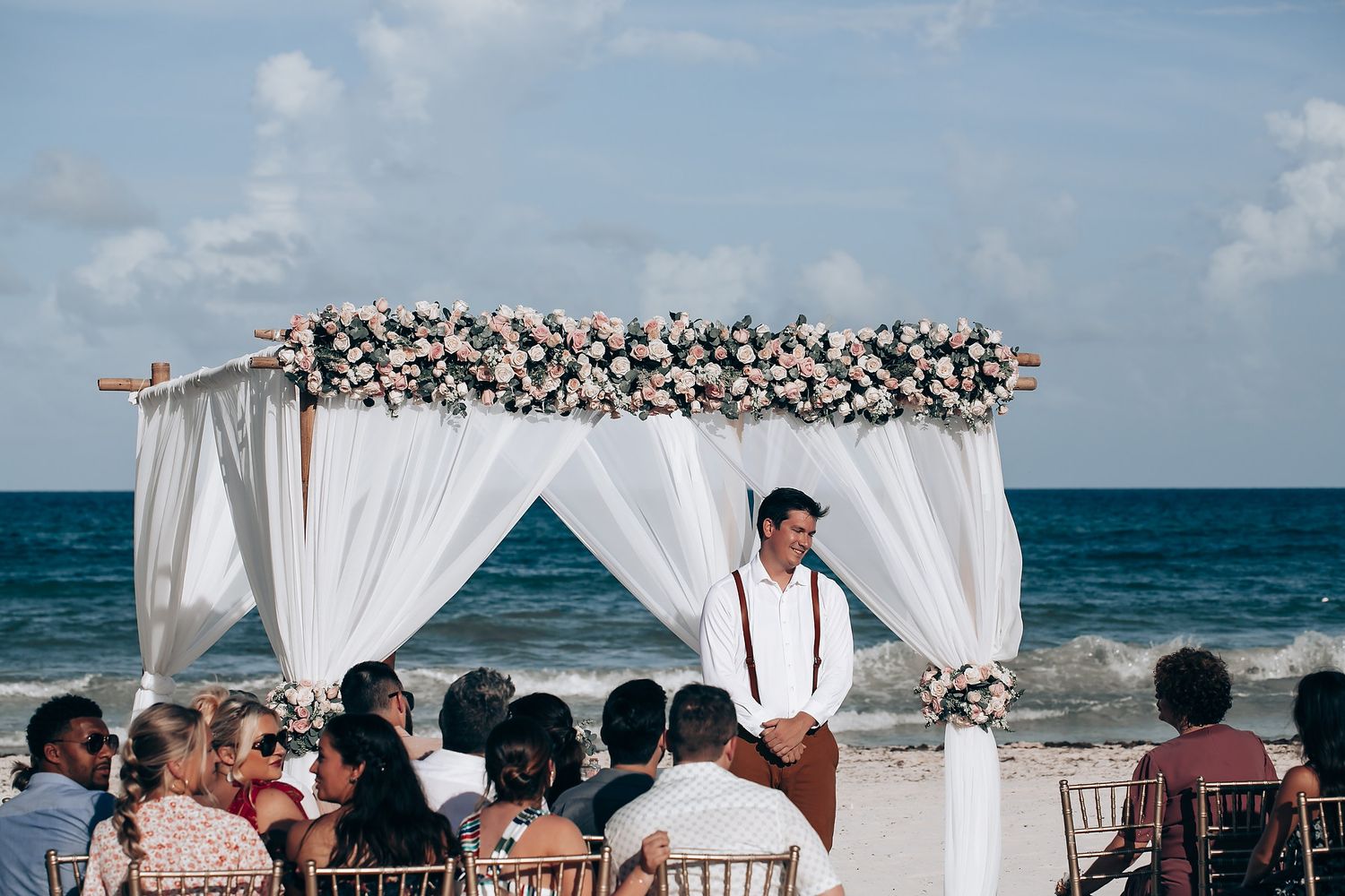 Beach wedding ceremony setup with white drapes and floral arch against ocean backdrop.