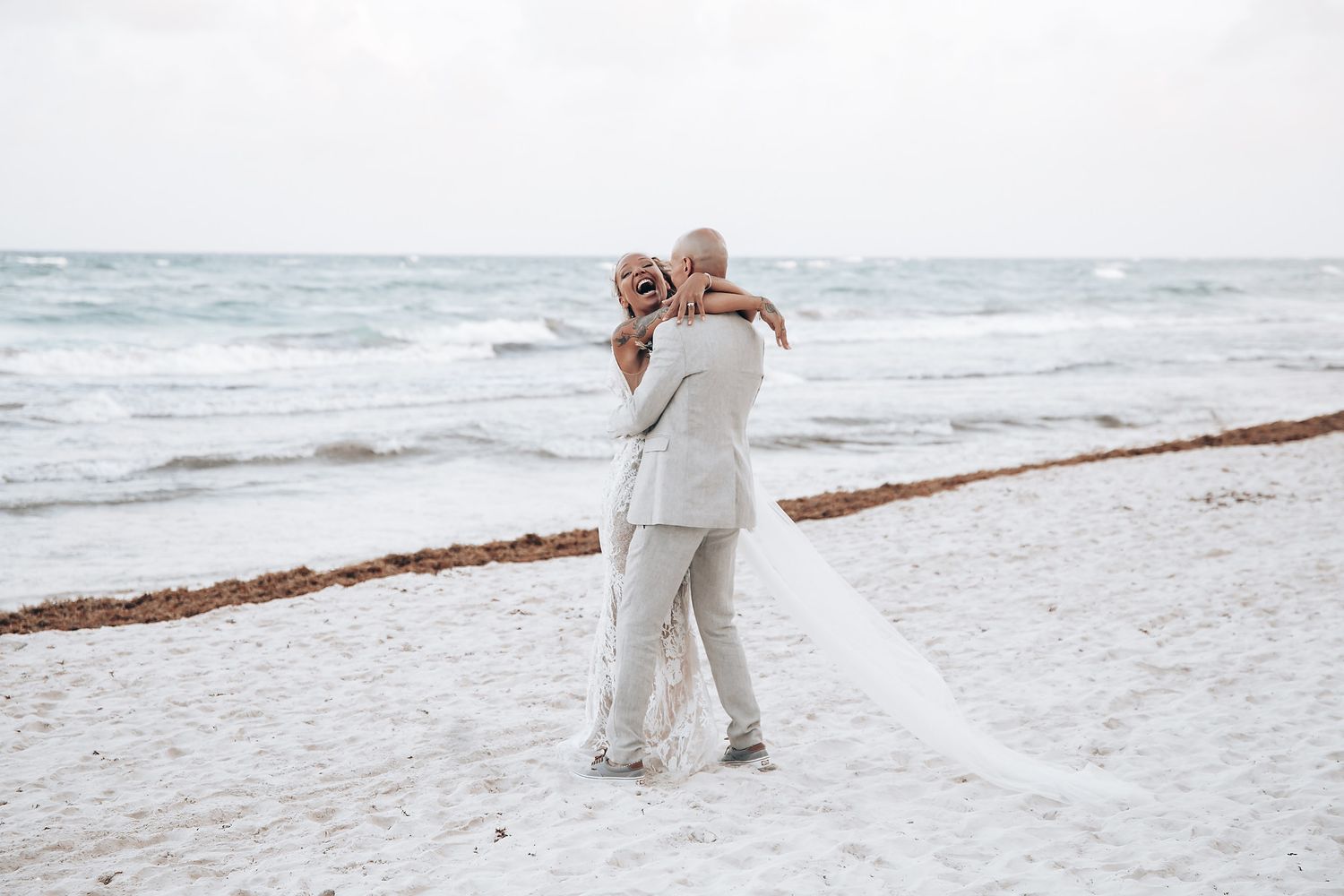 A newlywed couple embraces on a sandy beach with ocean waves in the background.