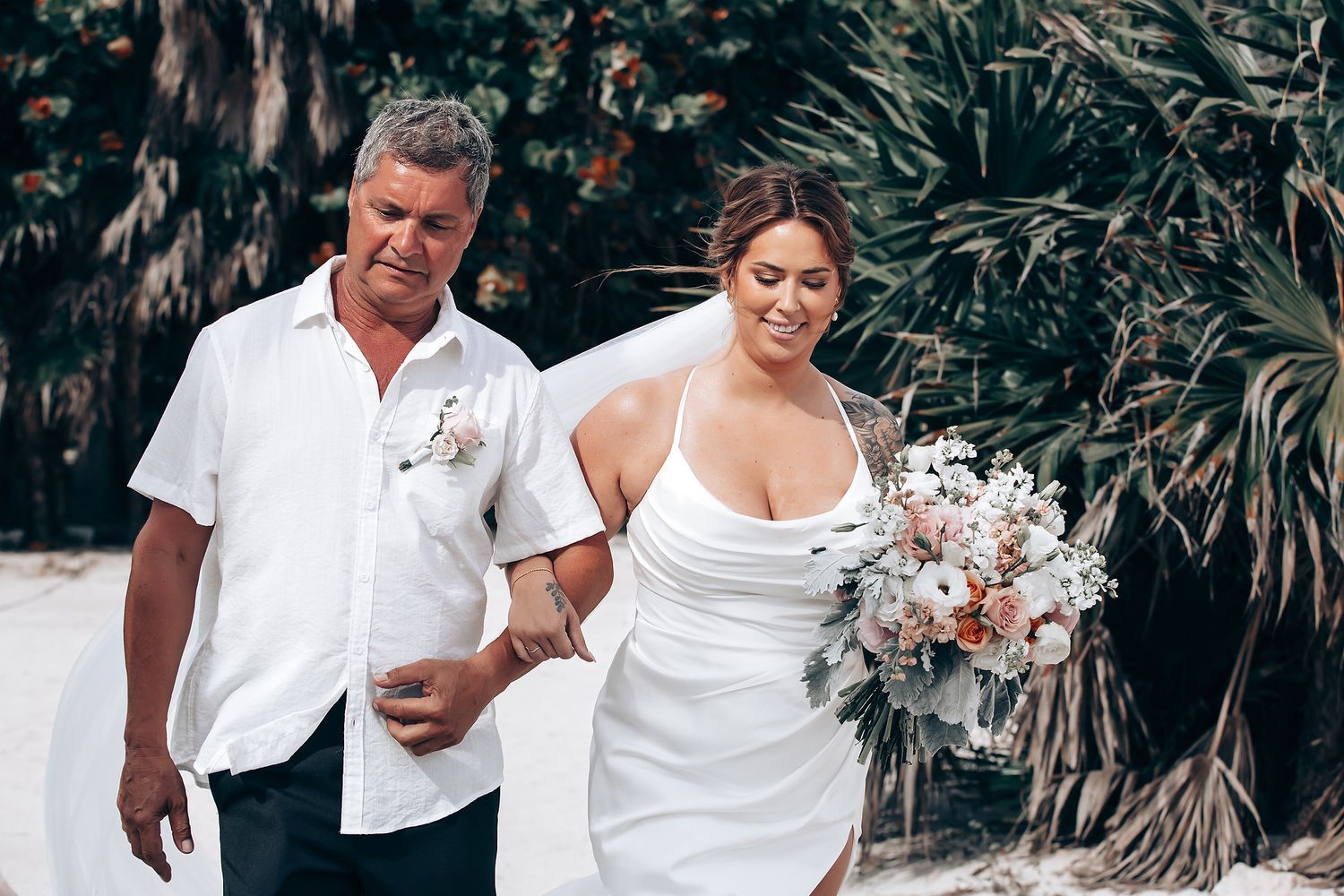 A couple in white outfits shares joyful moments in a tropical garden setting during a wedding celebration.