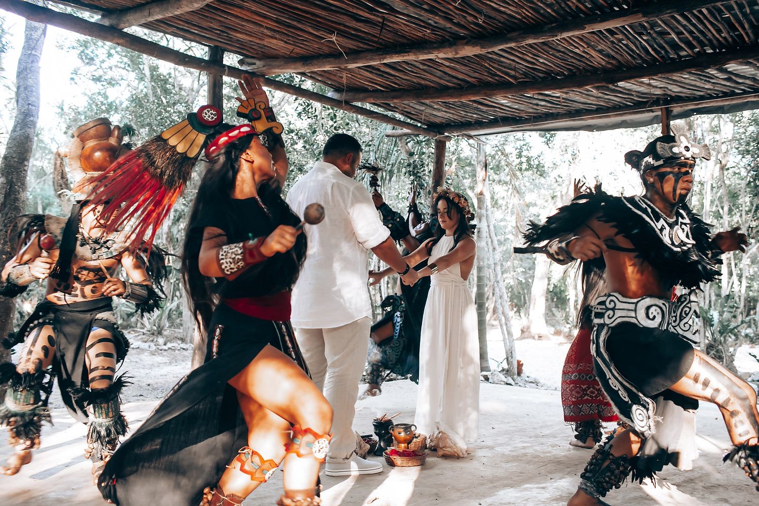 People engage in ceremonial activities under a rustic wooden shelter in nature.