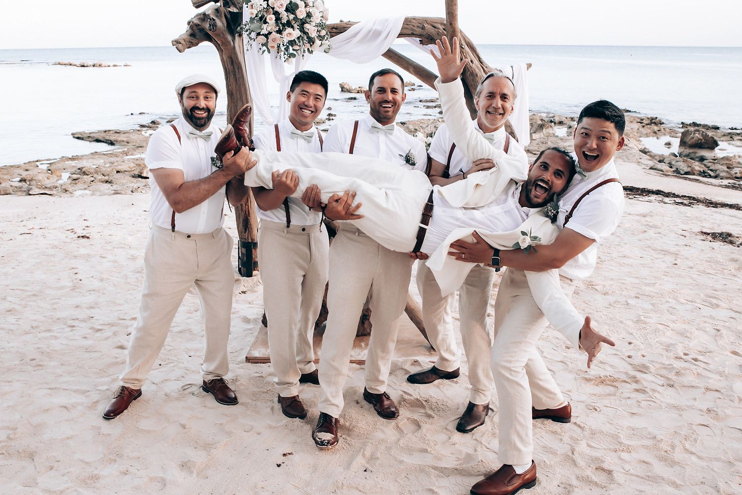 Wedding party in cream suits posing playfully on a sandy beach.