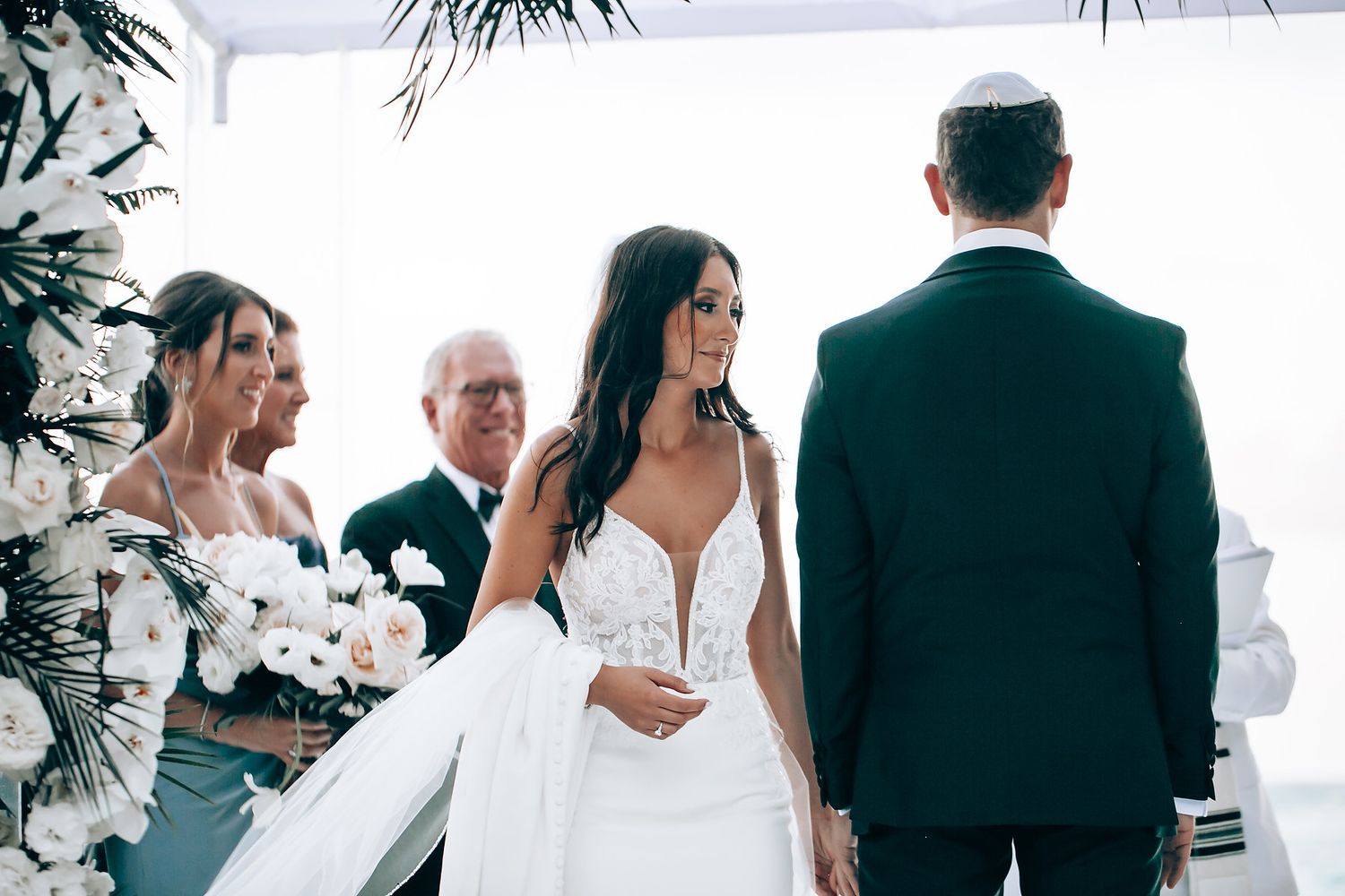 Wedding party members walk and interact during an outdoor celebration under a white tent.