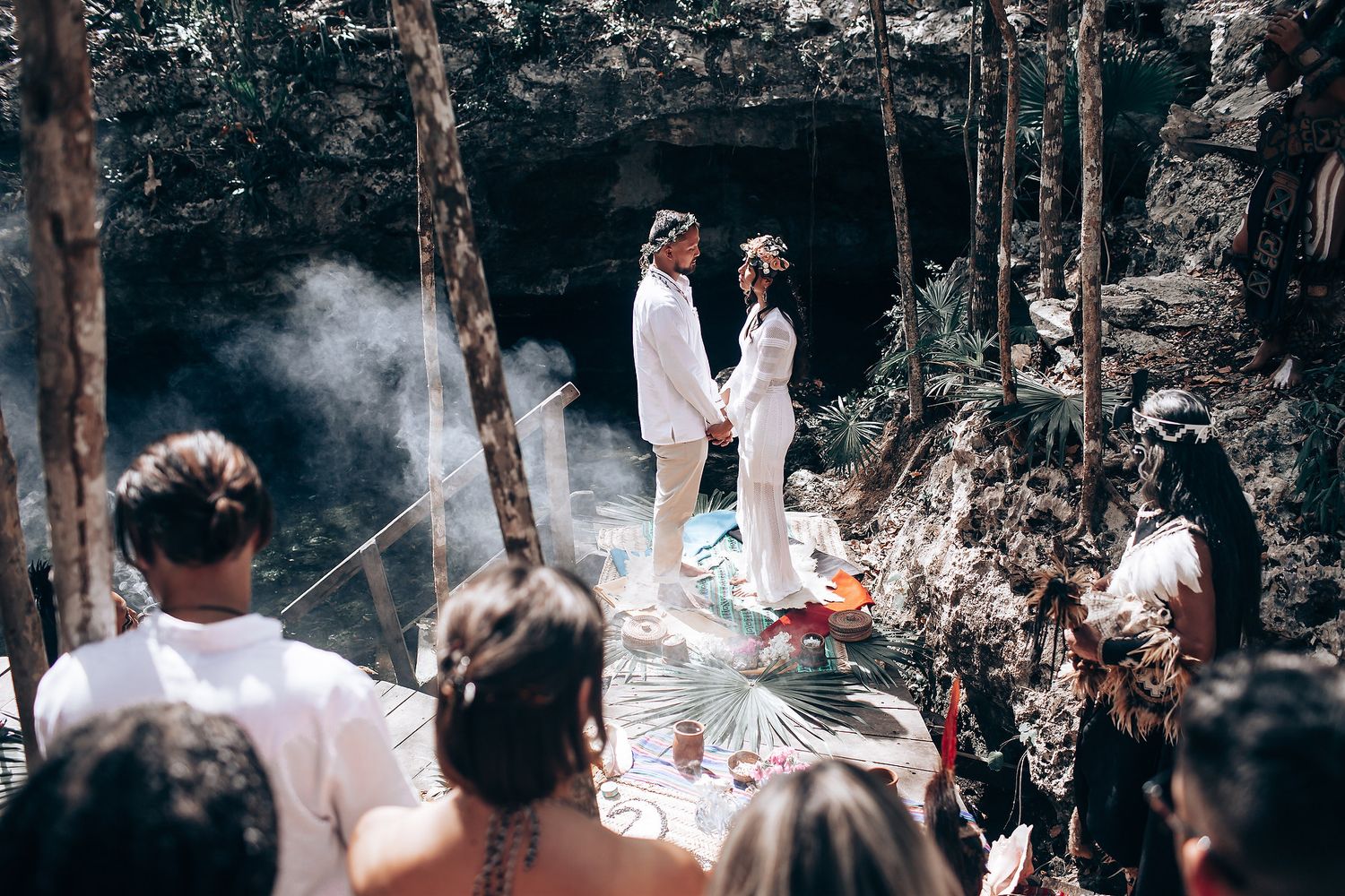 Ceremonial gathering in forest clearing with participants dressed in white performing traditional rituals.