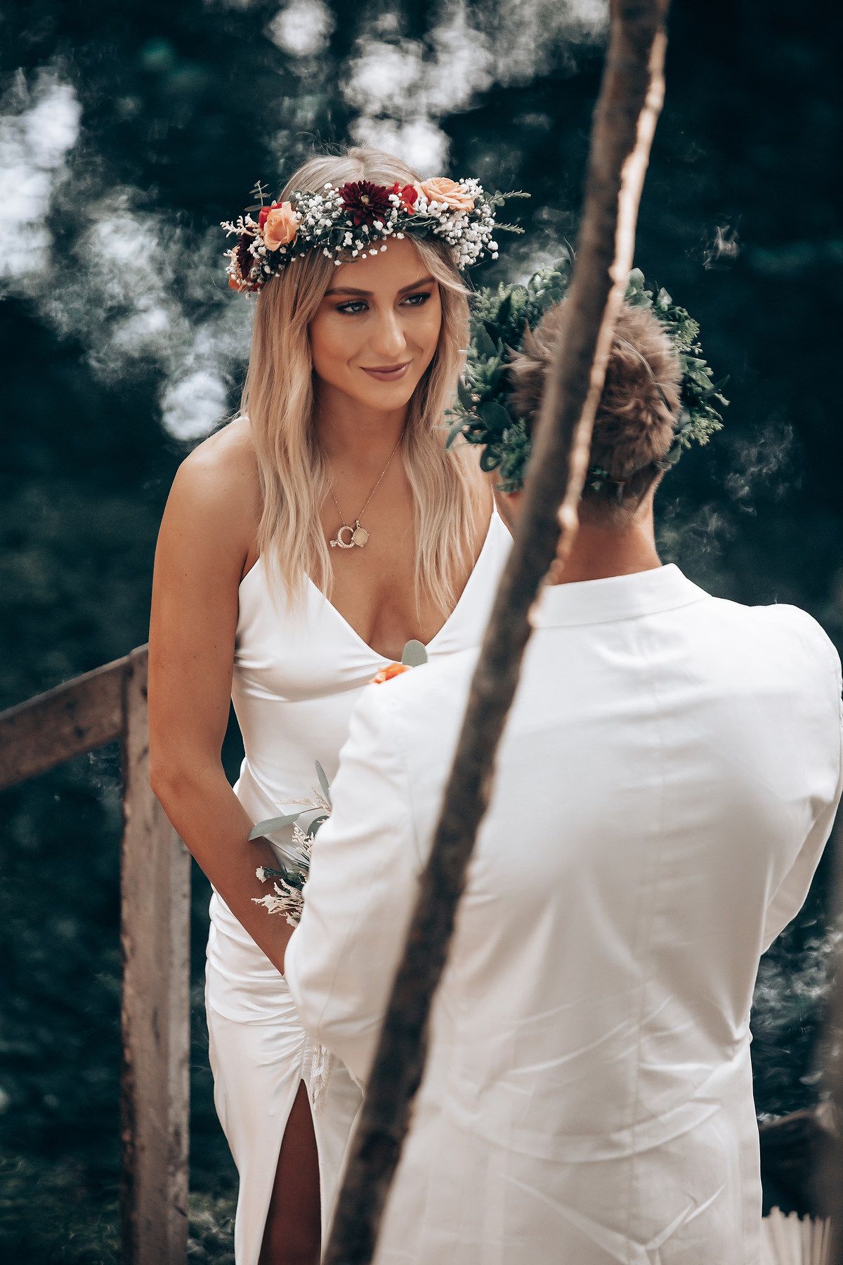 A bride wearing a white dress and floral crown poses in a moody outdoor setting.