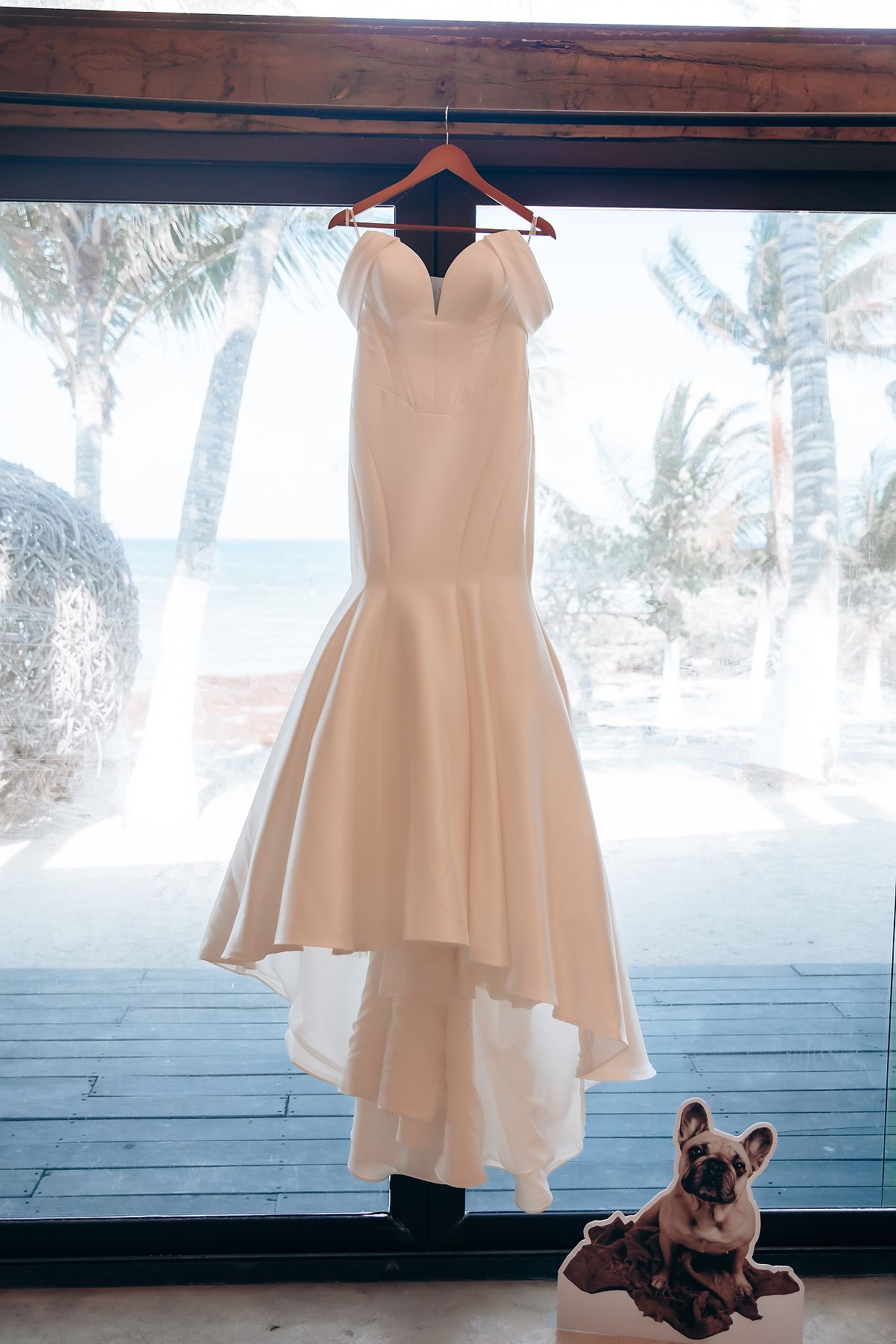 A light beige dress hangs elegantly against a blue wall while a French Bulldog sits curiously in the corner.
