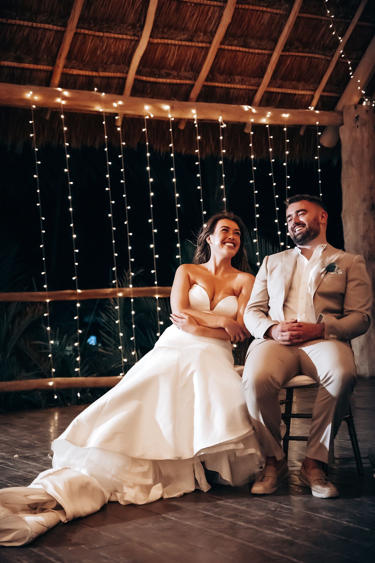 Newlywed couple sits on floor under string lights in rustic venue sharing intimate moments across four candid photos.