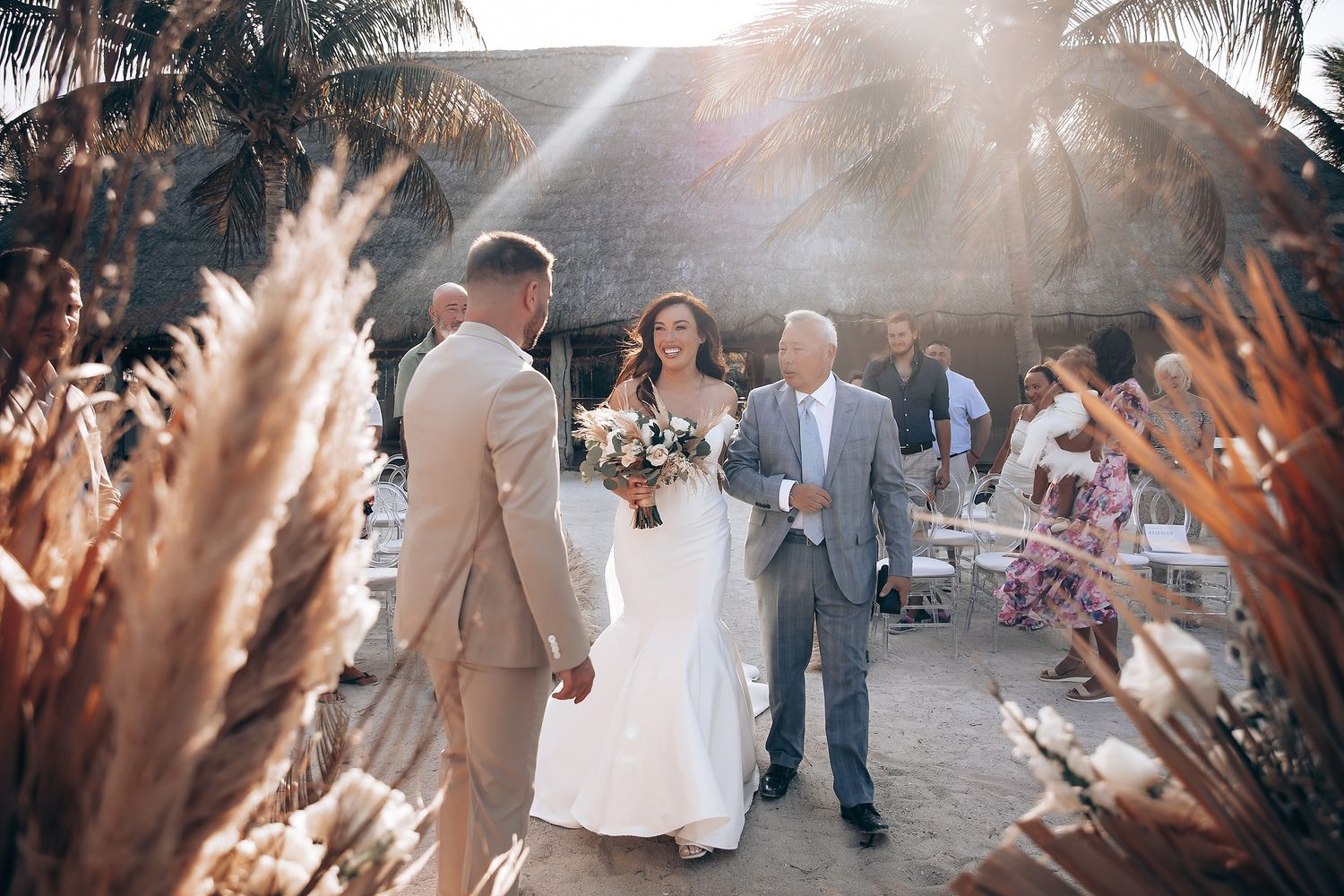Beachside wedding ceremony setup with pampas grass decorations and golden sunlight filtering through palm trees.