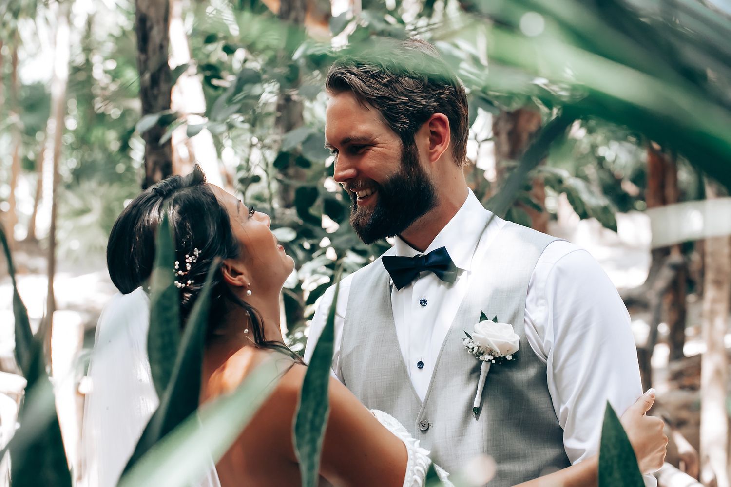 Wedding party members in formal attire gather beneath lush tropical foliage.