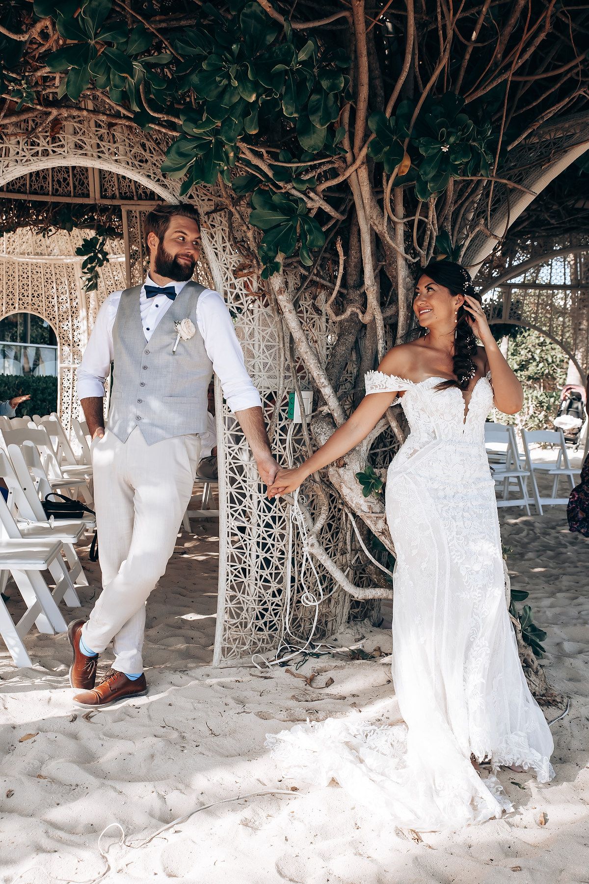 Wedding party in white attire poses playfully under a tree-covered beach pergola.