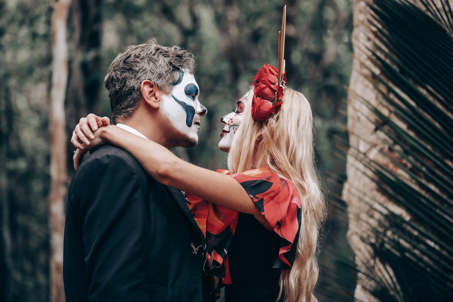 Couple in black and red outfits dance romantically in a forest setting with stone archways.