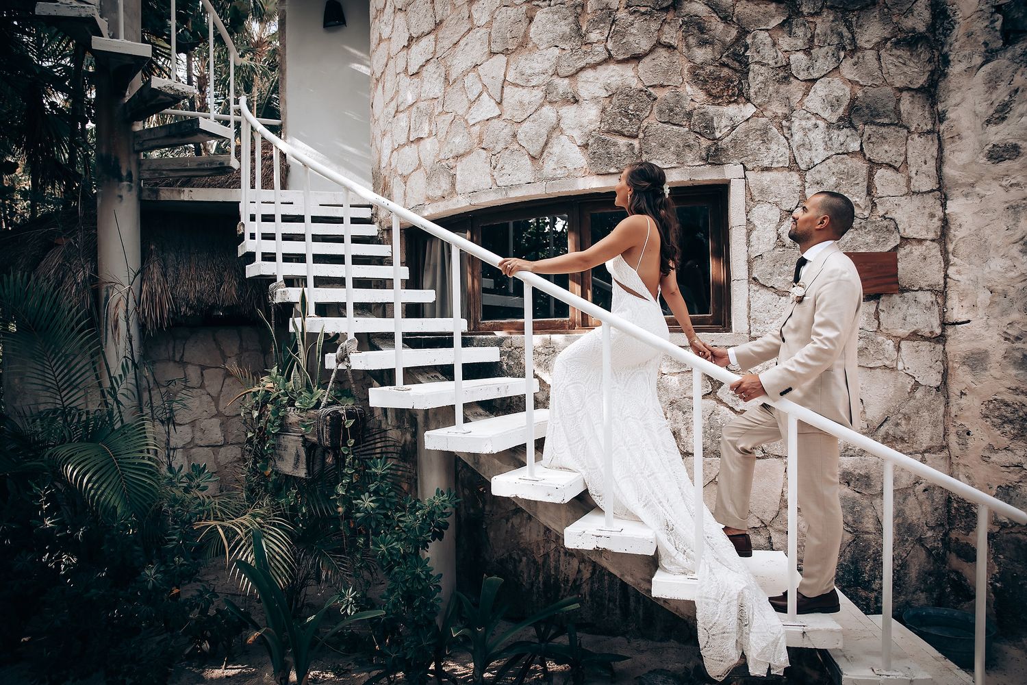 A couple in wedding attire poses on white spiral stairs against a stone wall in a romantic photoshoot sequence.