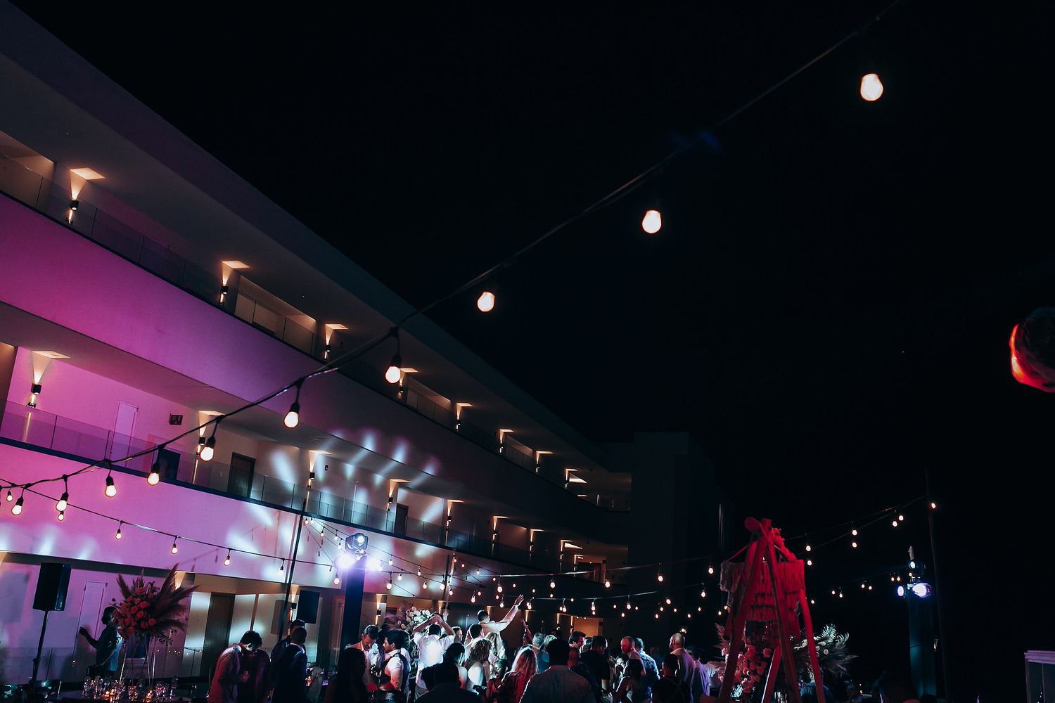 A dark event space illuminated by purple lighting and string lights with a crowd gathered inside.