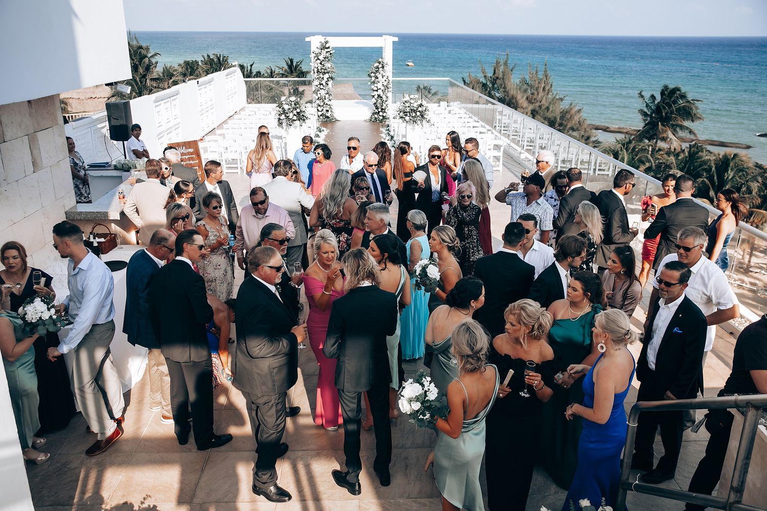 Guests gather at an oceanfront wedding ceremony with palm trees and blue skies in the background.