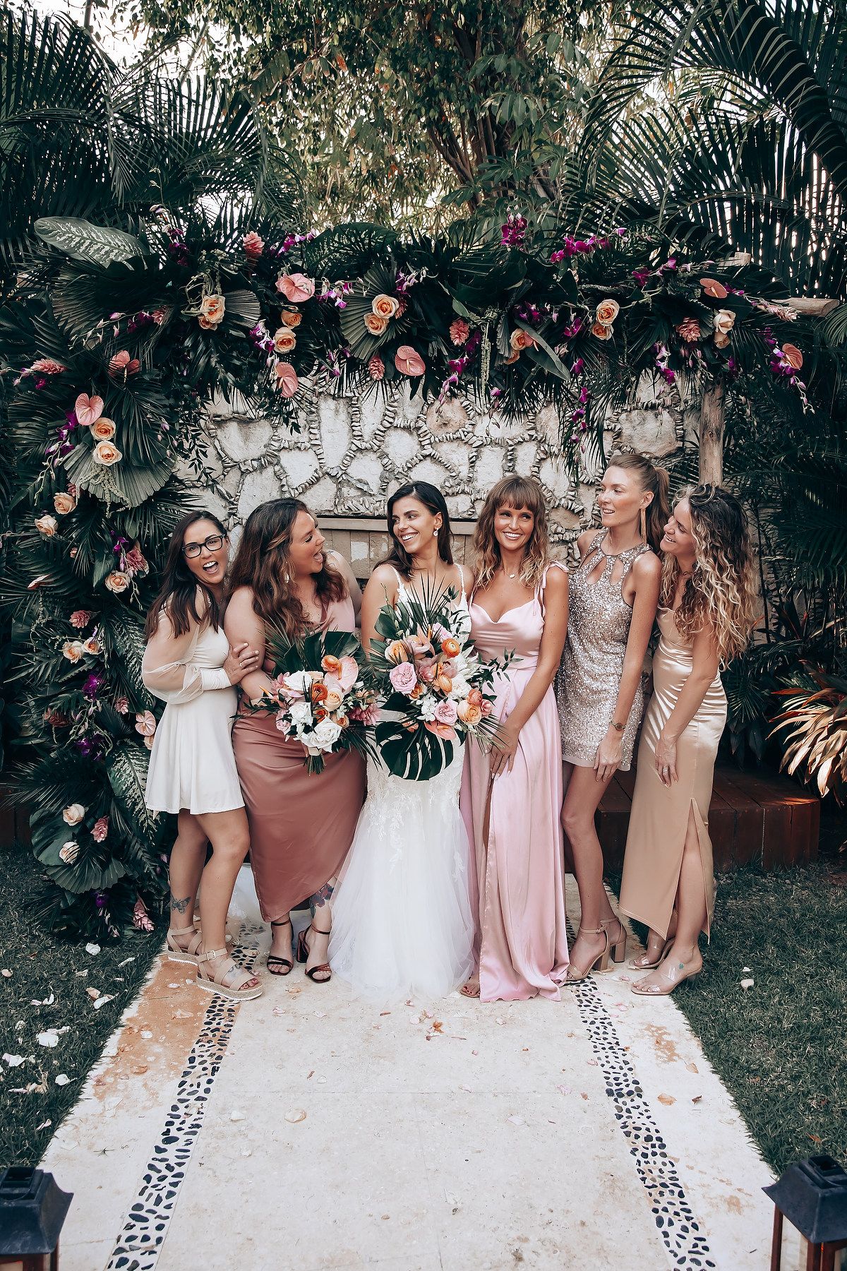 Bridesmaids in blush and neutral tones pose together on a white backdrop with floral decorations.
