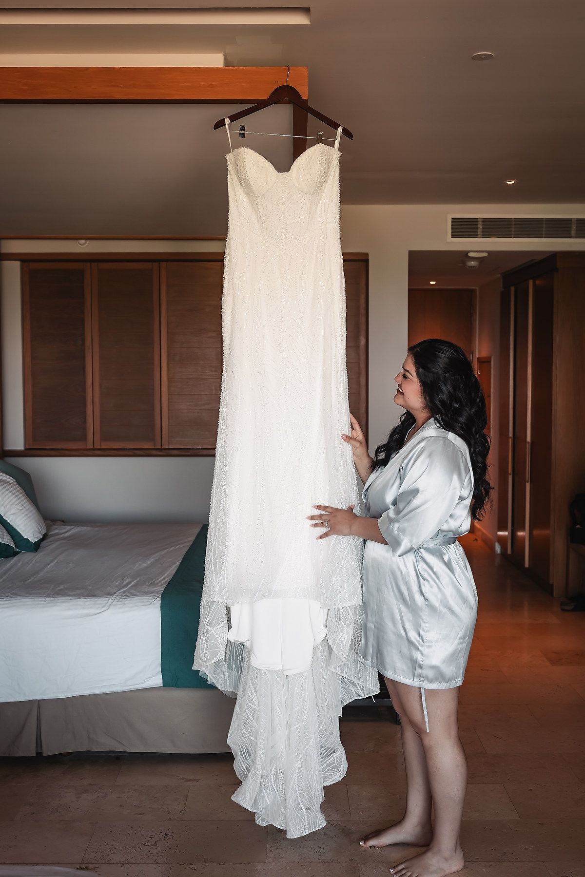 Bride in silver robe admiring her elegant white wedding gown hanging in a modern hotel room.