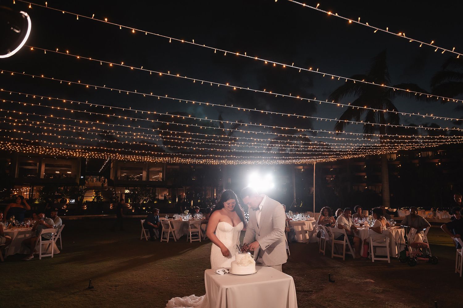 Bride and groom cutting wedding cake under romantic string lights at outdoor evening reception.