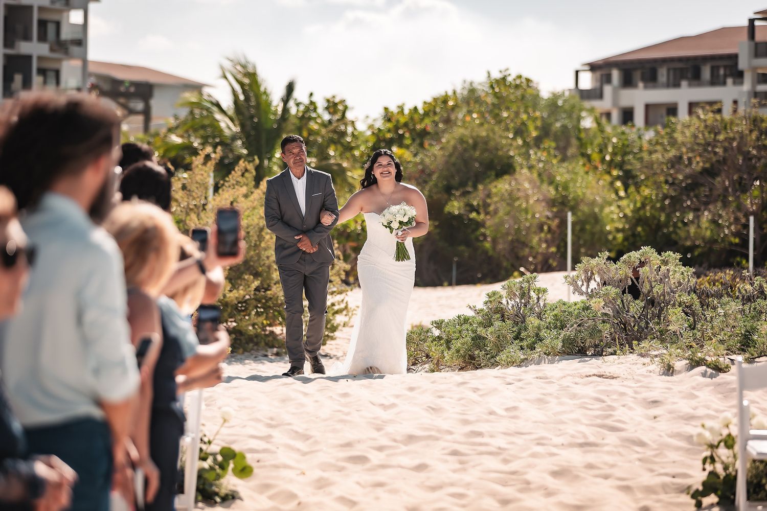 Bride and groom walking down sandy beach wedding aisle with guests watching and palm trees in background.