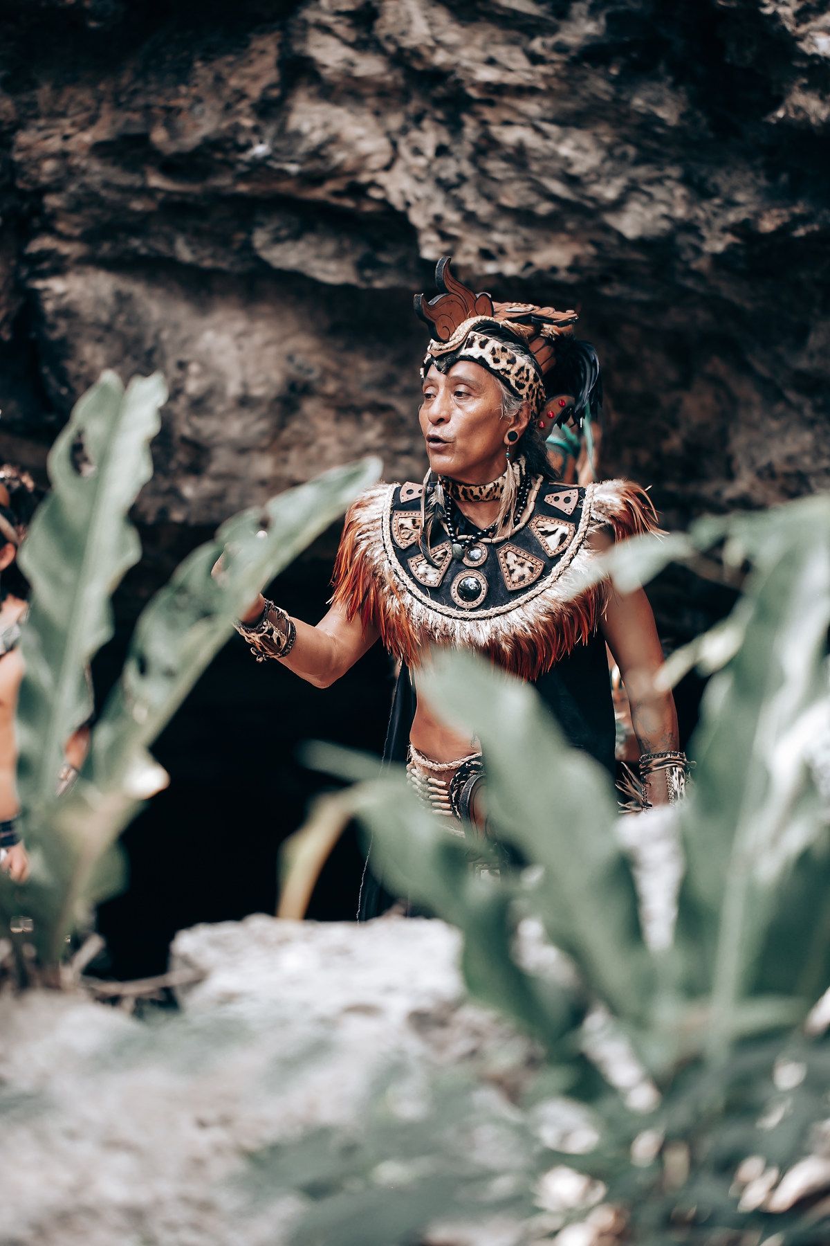 Man in traditional Mayan headdress and ornate necklace standing among tropical foliage near cave.