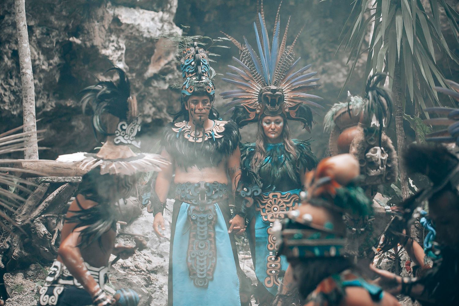 Two performers in stunning blue Mayan feathered headdresses and costumes in smoky cave setting.