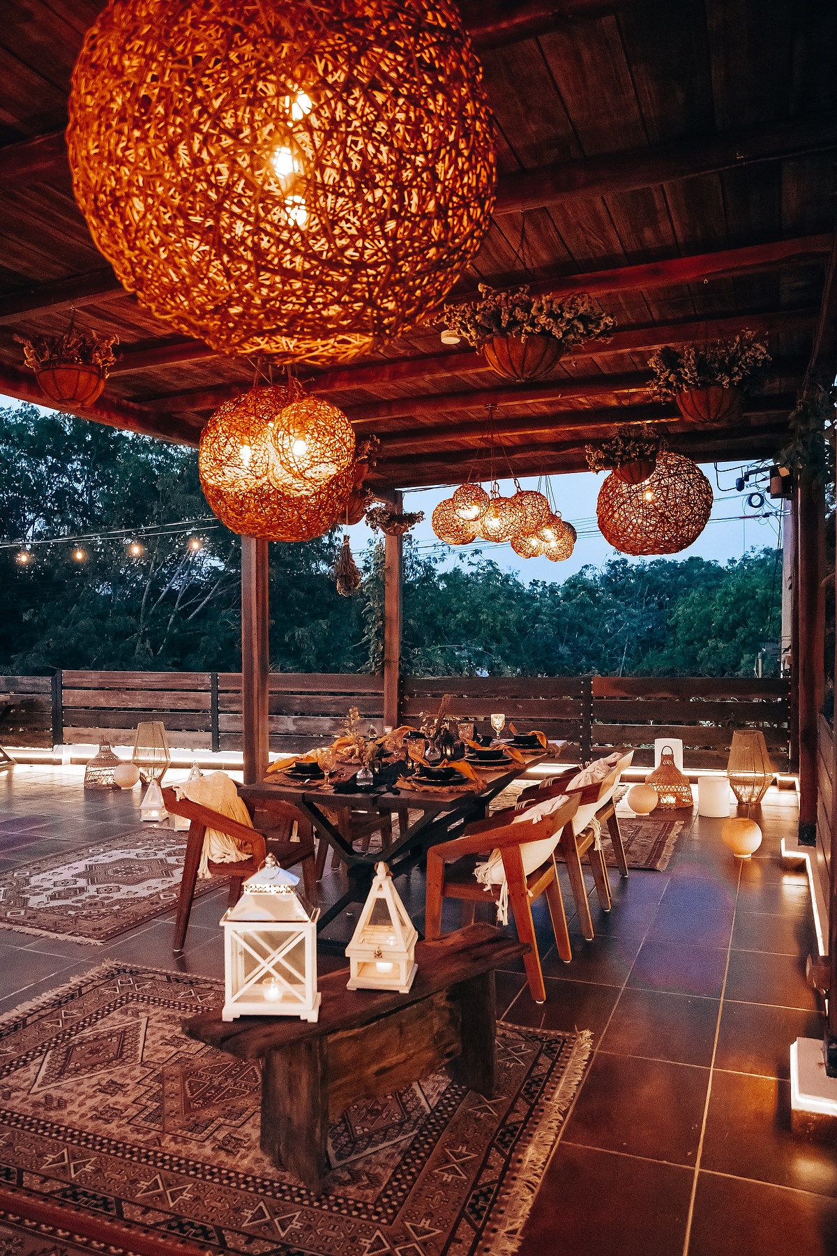 Rustic outdoor reception with large woven globe lights illuminating long wooden dining table at dusk.