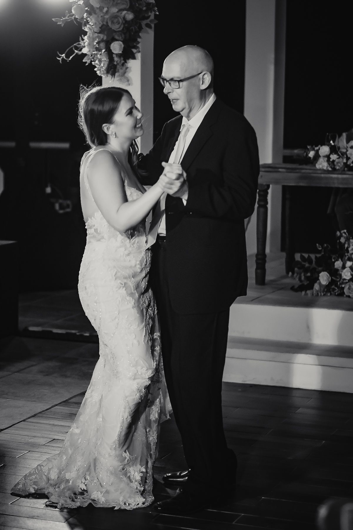 Black and white photo of a bride in lace gown sharing a father-daughter dance at an elegant wedding reception.