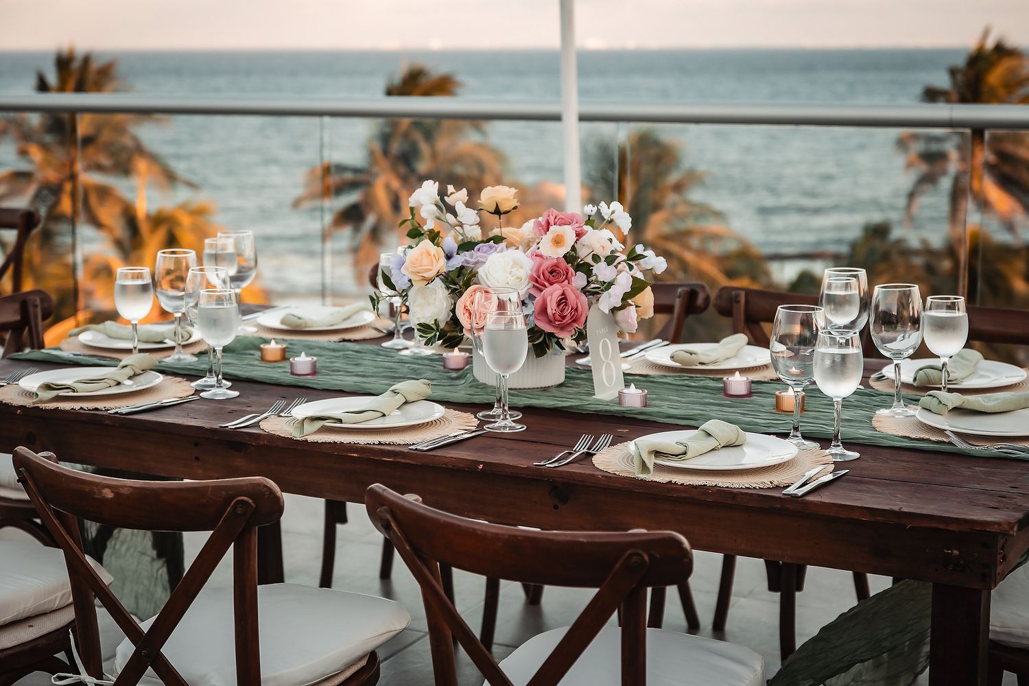 Elegant outdoor beachfront wedding reception table with floral centerpiece, wine glasses, and ocean view at sunset.