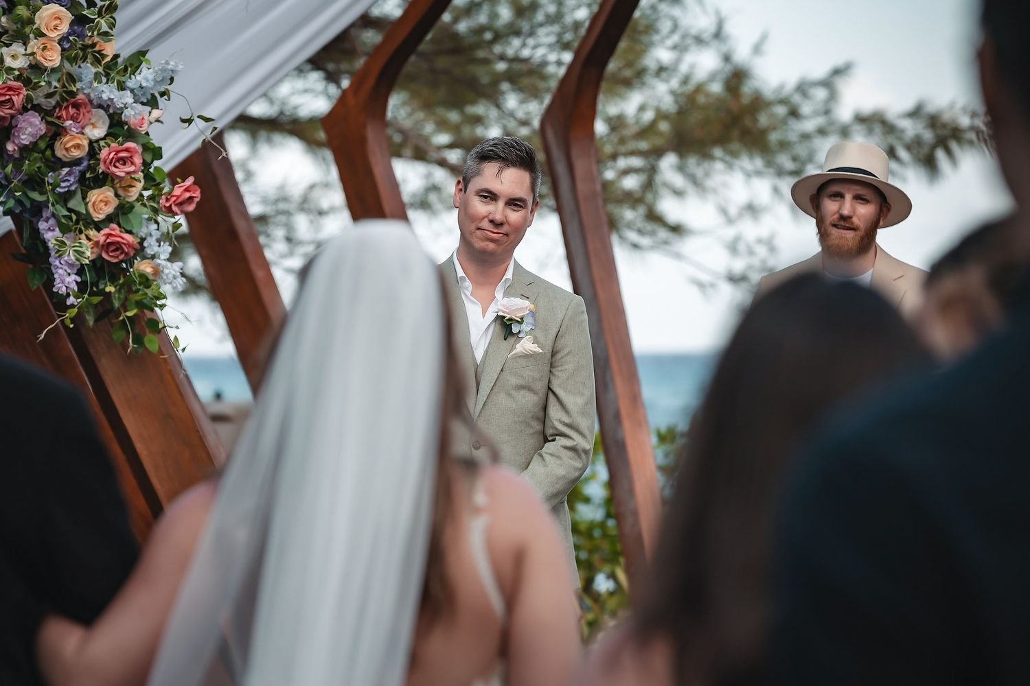 Groom in beige suit stands at outdoor wedding altar with floral arrangements, awaiting bride.