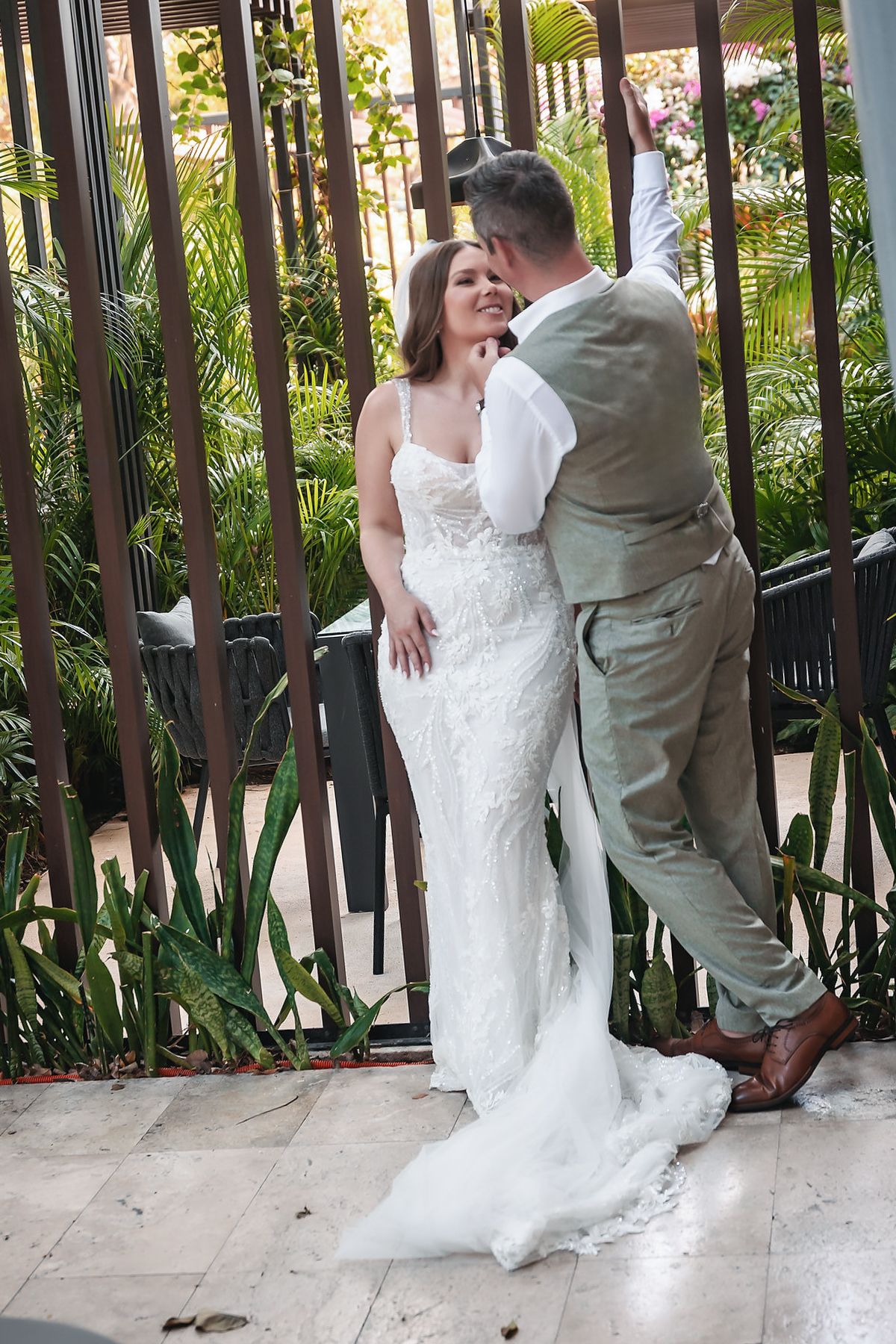 A bride in a white lace gown and groom in a gray suit share a romantic moment by decorative iron gates surrounded by tropical greenery.