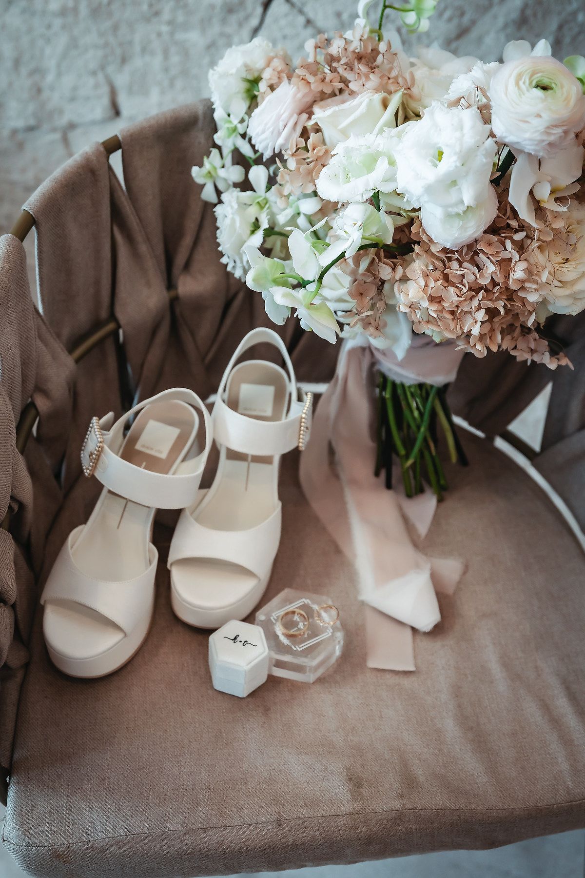 Elegant bridal flat lay featuring cream platform sandals, wedding rings in a clear box, and a soft pink and white floral bouquet on a chair.