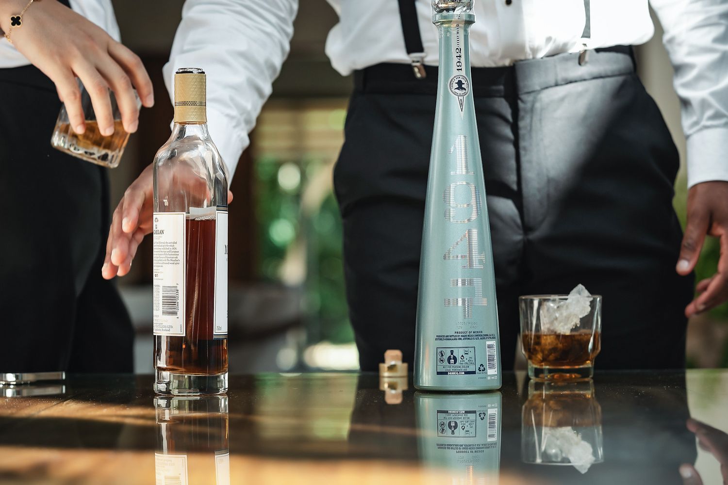 Two bartenders pouring premium whiskey and tequila bottles on a reflective wooden bar counter.