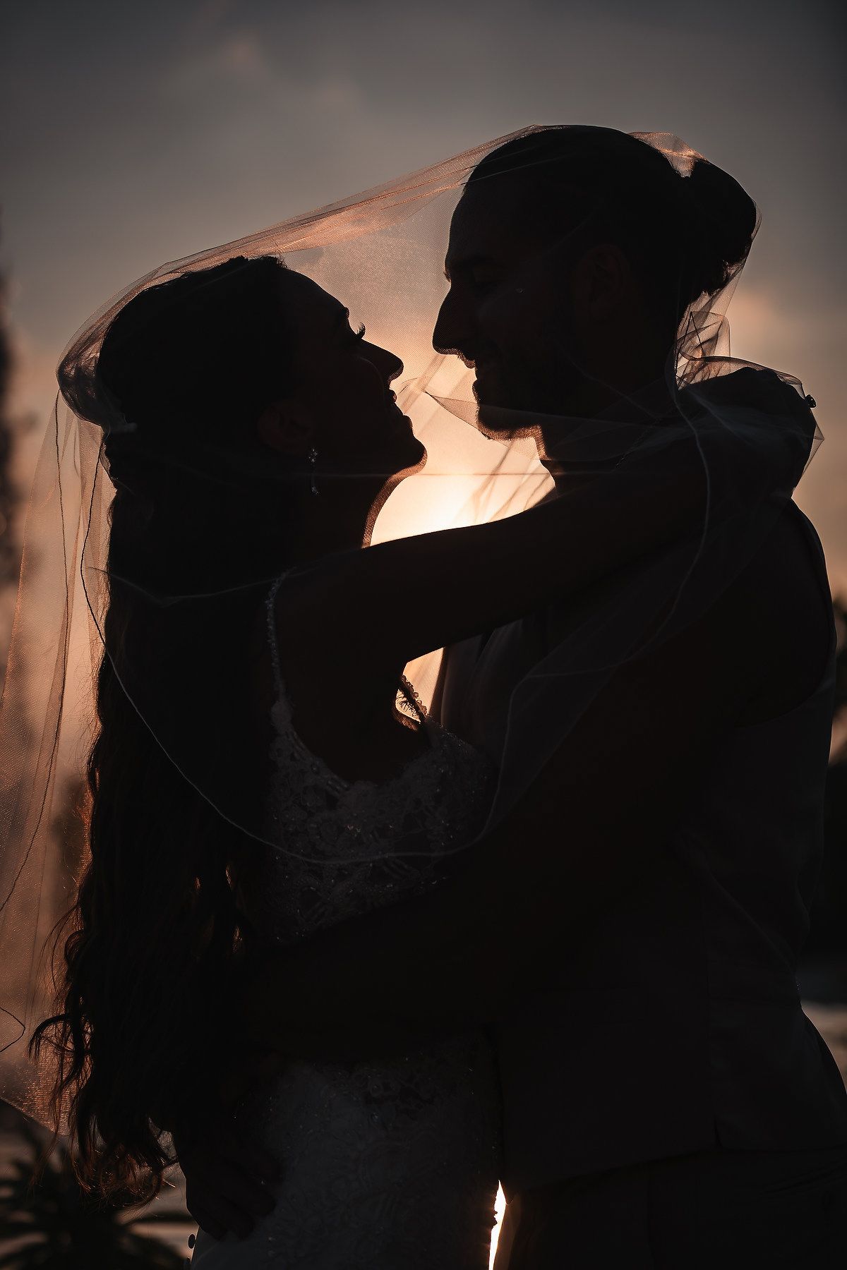 Silhouette of a bride and groom embracing under a veil at sunset, sharing a romantic moment.